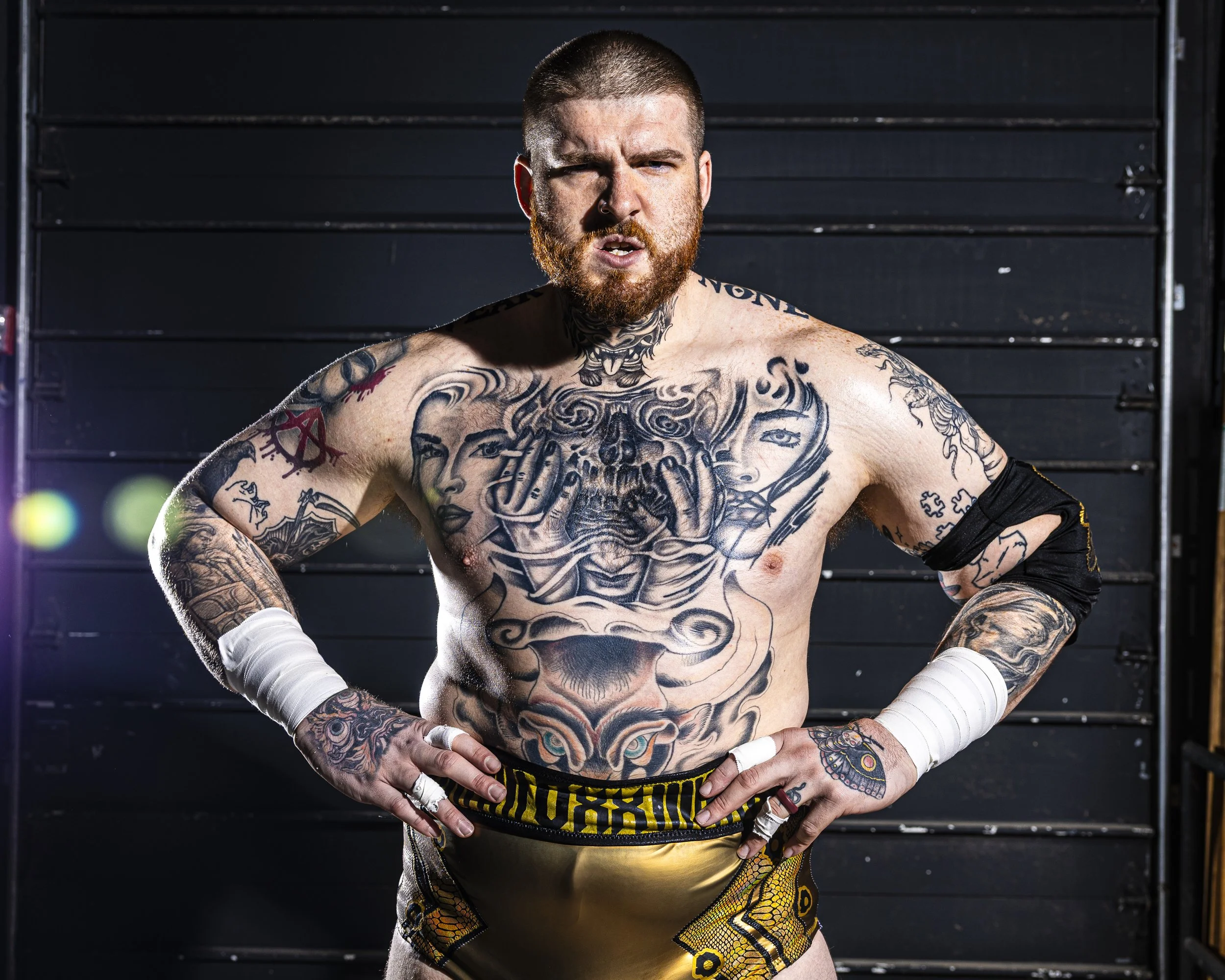 A muscular, tattooed man standing with his hands on his hips and a confident expression, wearing boxing shorts and white hand wraps, in front of a black background with a gray metal structure.