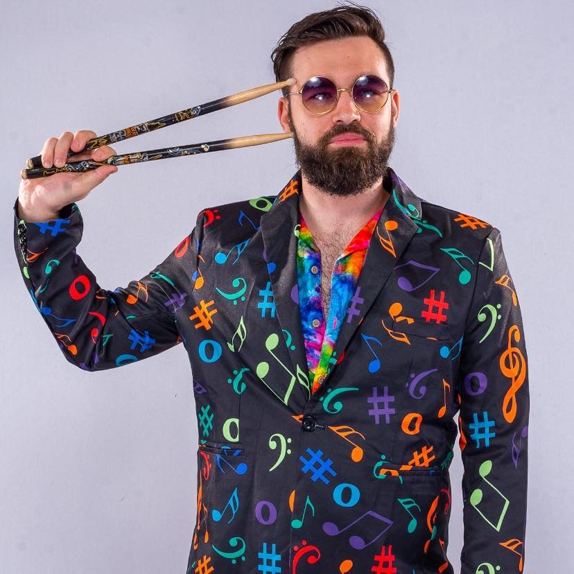 A man with a beard and sunglasses wearing a black suit with colorful musical note patterns, holding drumsticks to his temple, and sporting a rainbow tie.