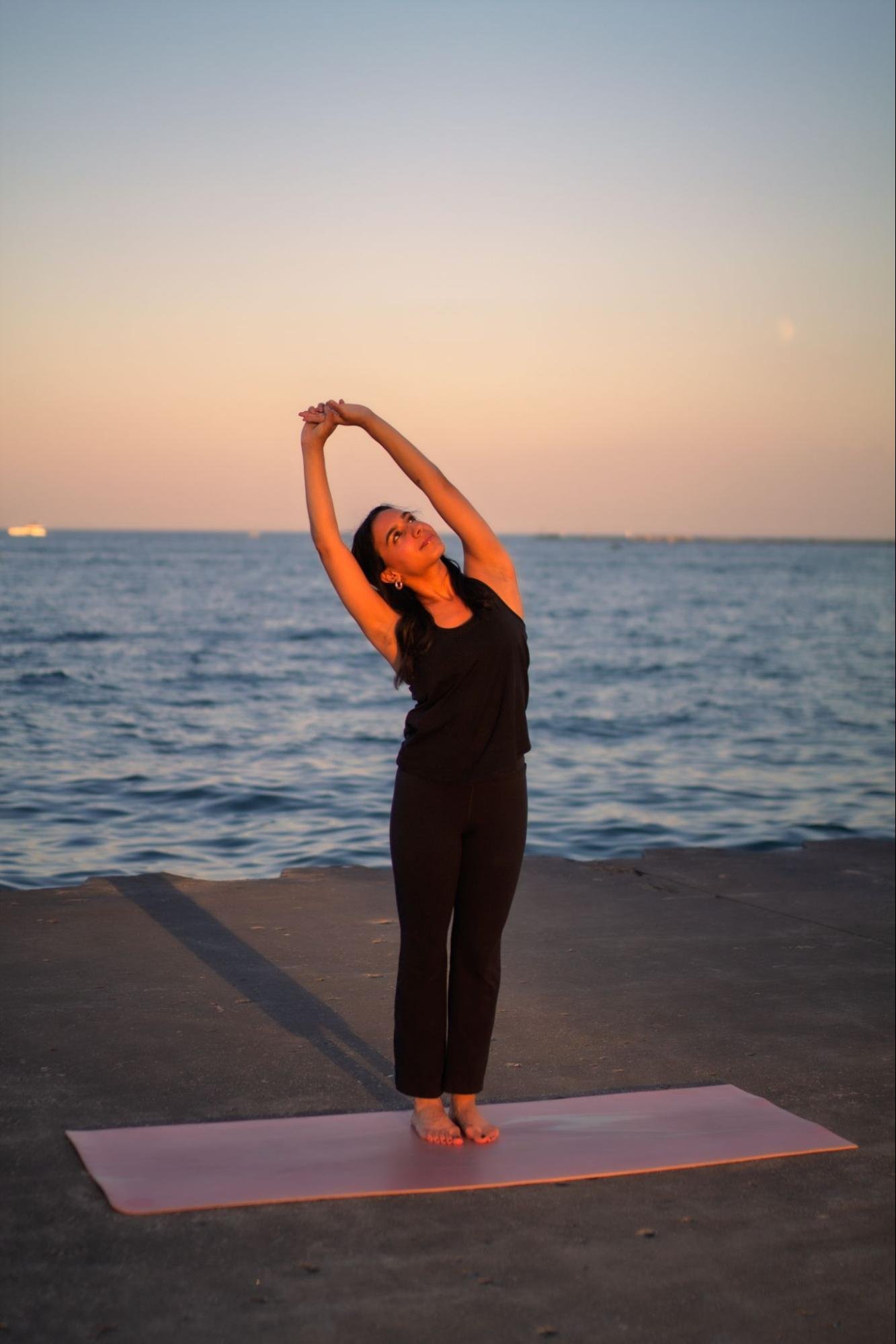 A woman practicing yoga on a mat near the water during sunset, standing in a stretch pose with arms raised above her head