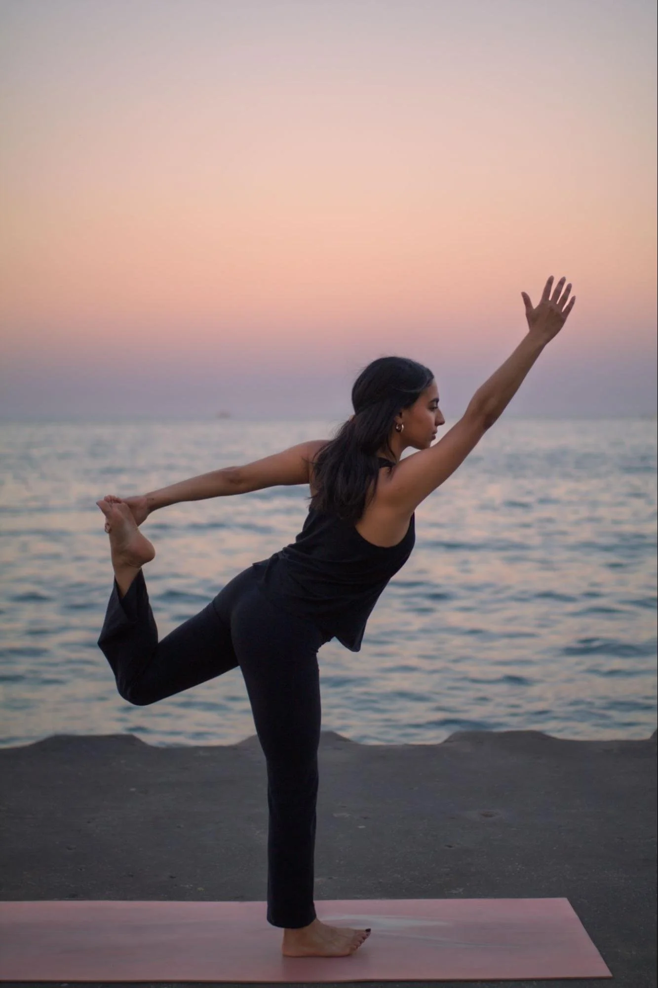 A woman practicing yoga outdoors on a beach at sunset, standing on one leg with her other leg bent behind her and held by her hand, forming a graceful pose with one arm extended forward and the other holding her foot.