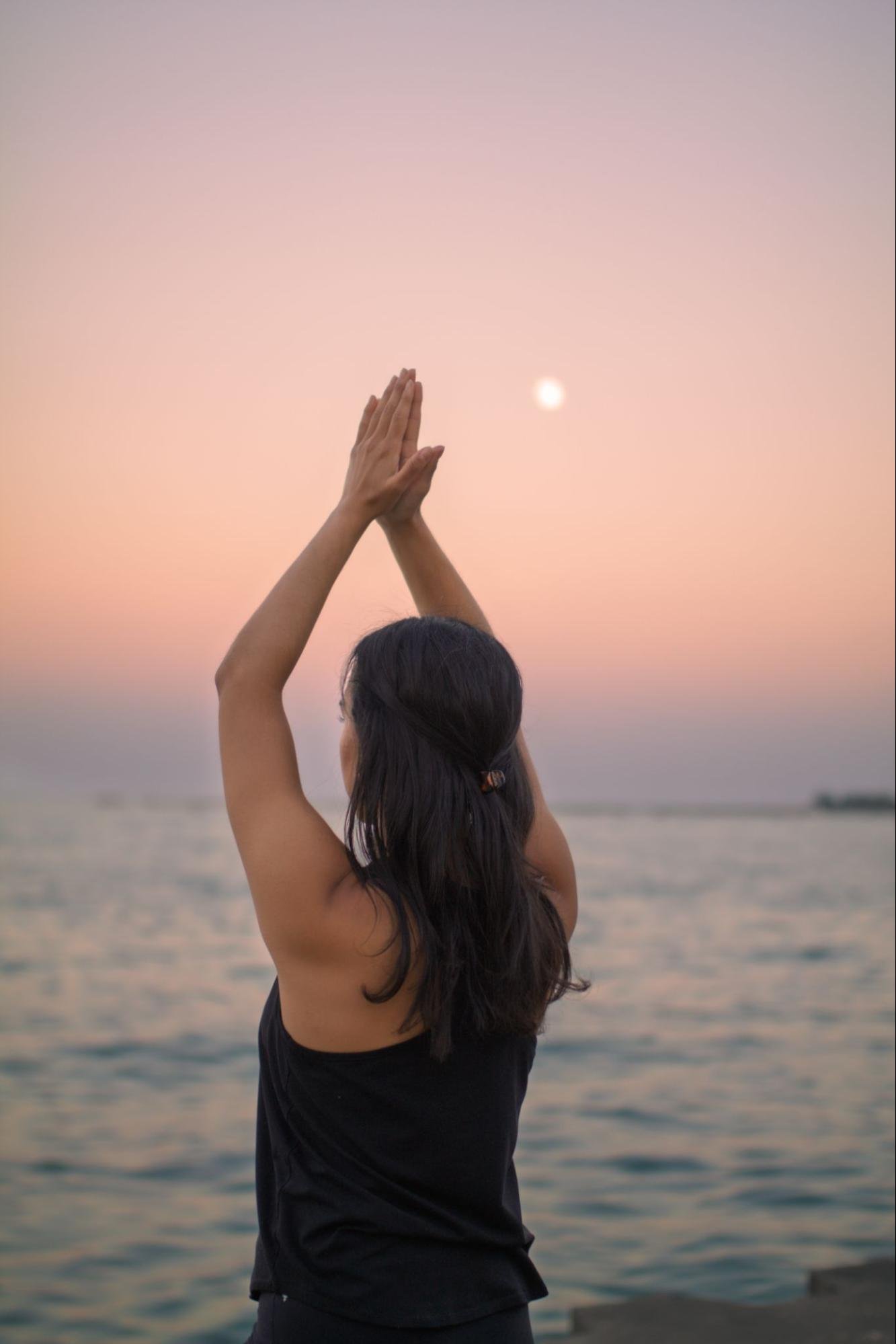 A person with long dark hair practicing yoga or meditation outdoors near water at sunset, with hands together in prayer pose and the sun low in the sky.