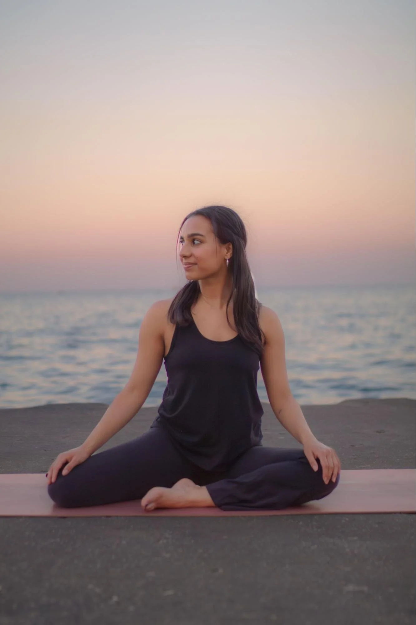 A woman practicing yoga on a mat outdoors near a body of water at sunset, sitting in a cross-legged pose with hands resting on her knees, smiling softly and looking to the side.