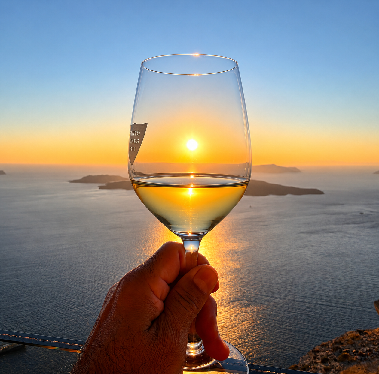 Glass of white wine held up against a Santorini sunset over the caldera, with a Santo Wines label visible.