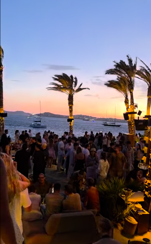 Crowd gathered at sunset at Scorpios beach club in Mykonos, Greece, with palm trees and boats on the water.