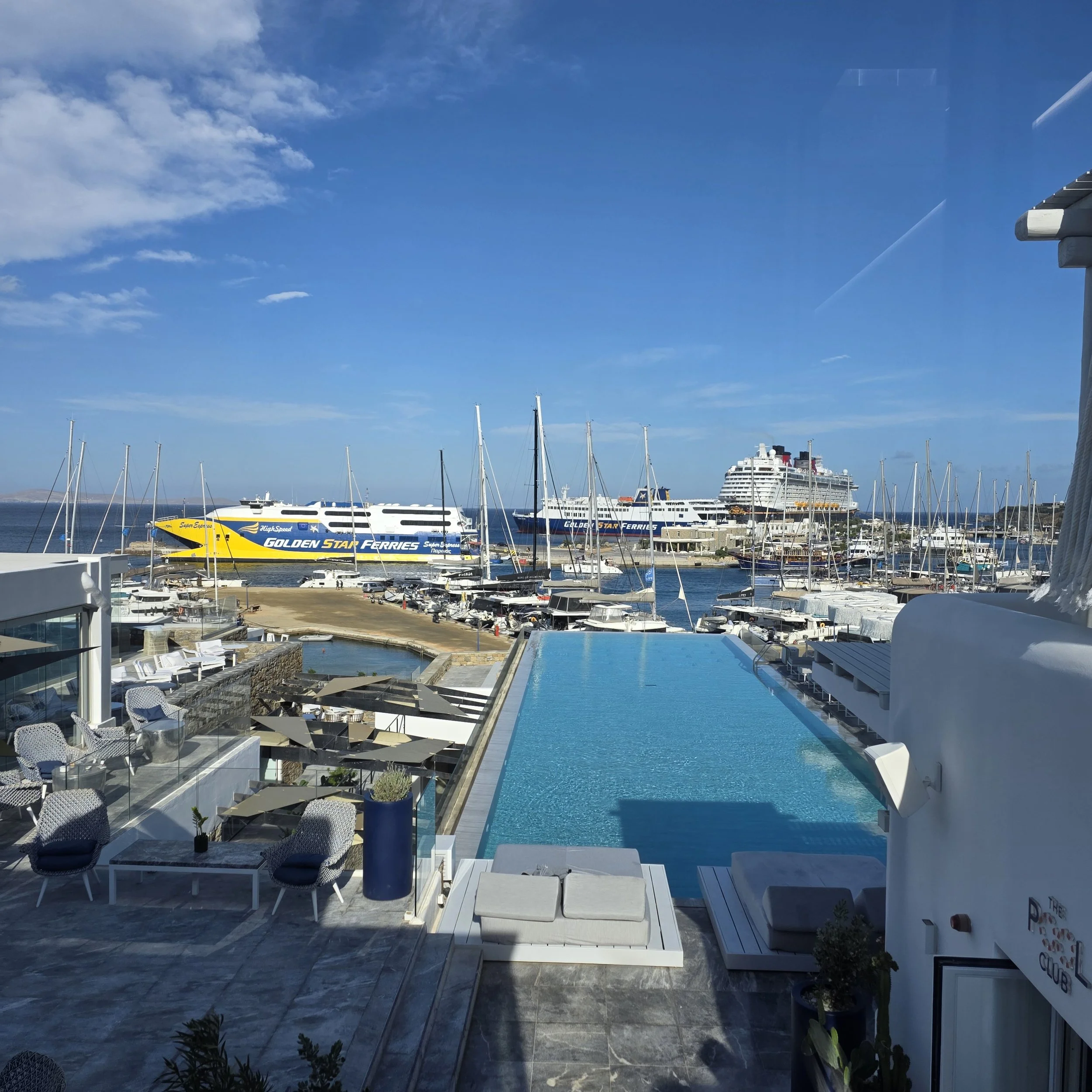 Infinity pool at Mykonos Riviera Hotel overlooking the marina, with sailboats, ferries, and a cruise ship in the background.