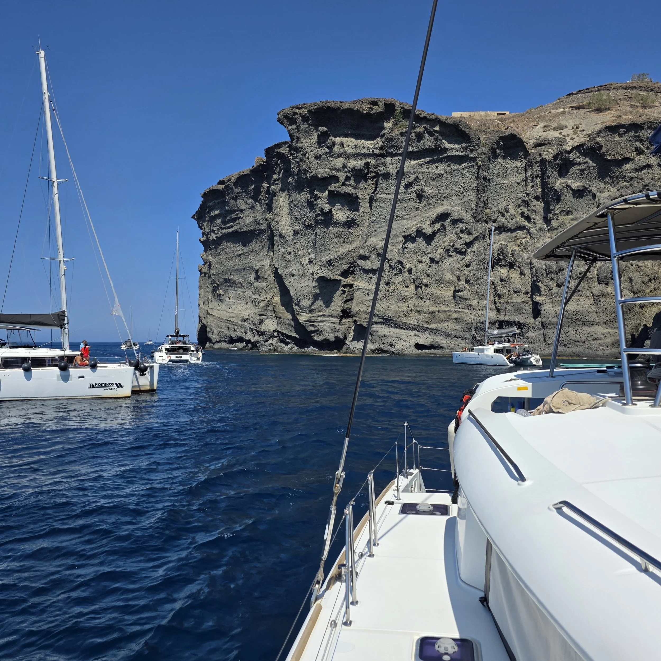 Sailboats anchored near volcanic cliffs off the coast of Santorini, Greece, during a catamaran sunset tour.