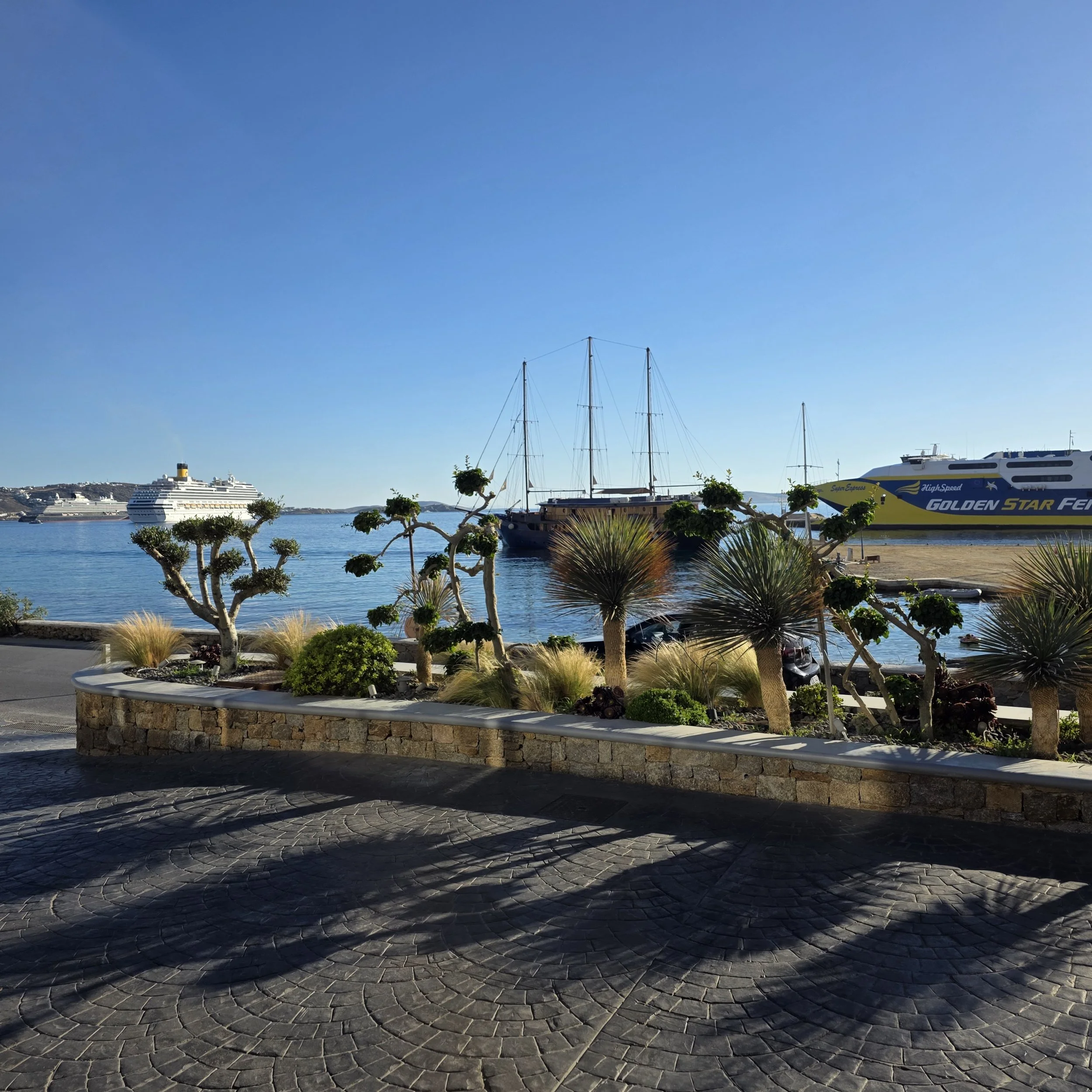 Mykonos harbor with a cruise ship, sailboat, and ferry docked, framed by sculpted trees and stone landscaping.
