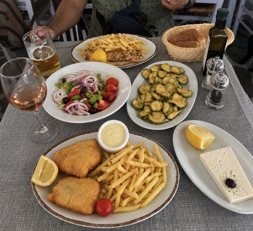 Greek taverna table with fried fish, fries, Greek salad, fried zucchini, feta, and wine at Balconi Tou Aki, Paros.