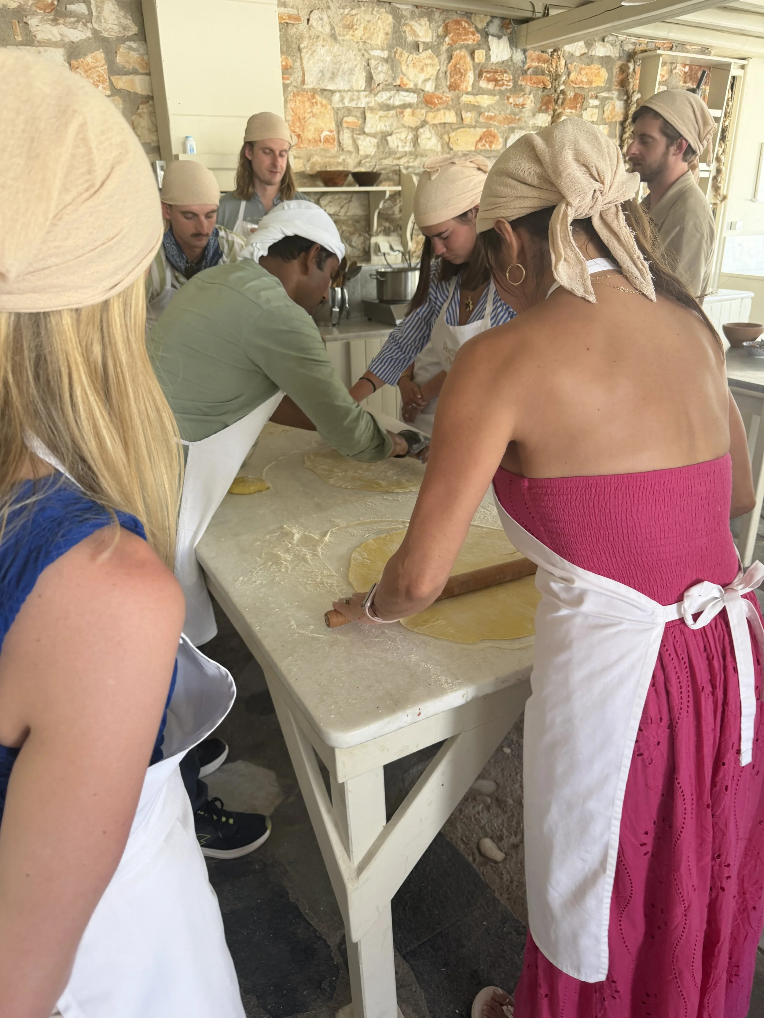 Group hand-rolling dough at an outdoor Greek cooking class table on Paros, with a stone wall kitchen in the background.