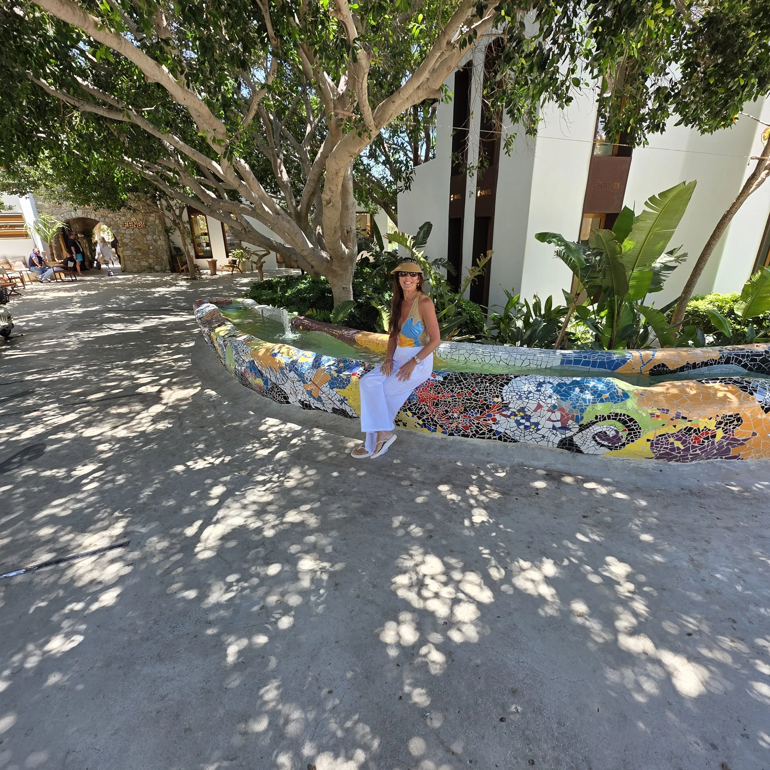Woman sitting on a mosaic-tiled fountain bench at Nammos Village in Mykonos, Greece, with Fendi and Chanel storefronts in the background.