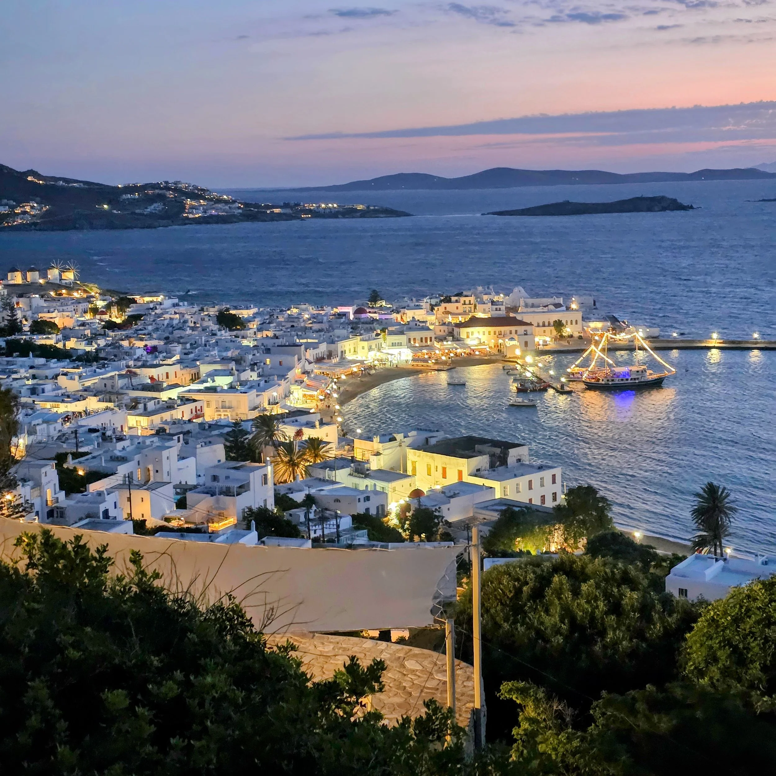 Mykonos harbor at dusk with whitewashed buildings and boats lit up at night