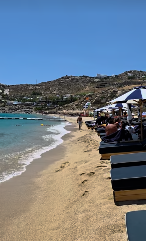 Sandy beach walkway lined with sun loungers and umbrellas at Super Paradise beach club in Mykonos, Greece.