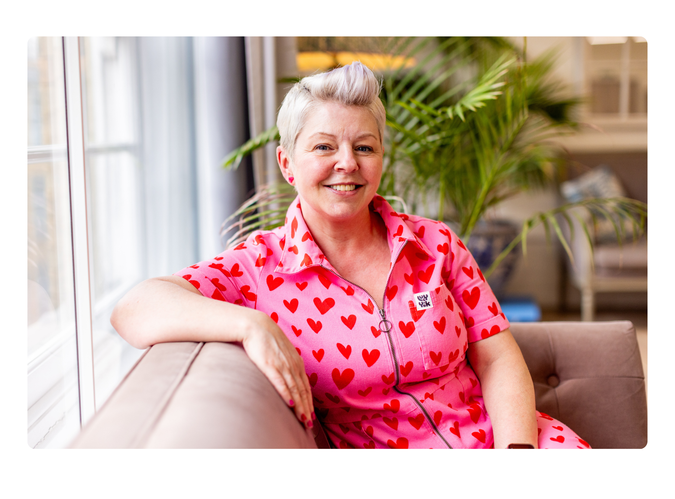 Picture of Jenny Martin wearing a pink jumpsuit, sitting in an armchair, with plants behind