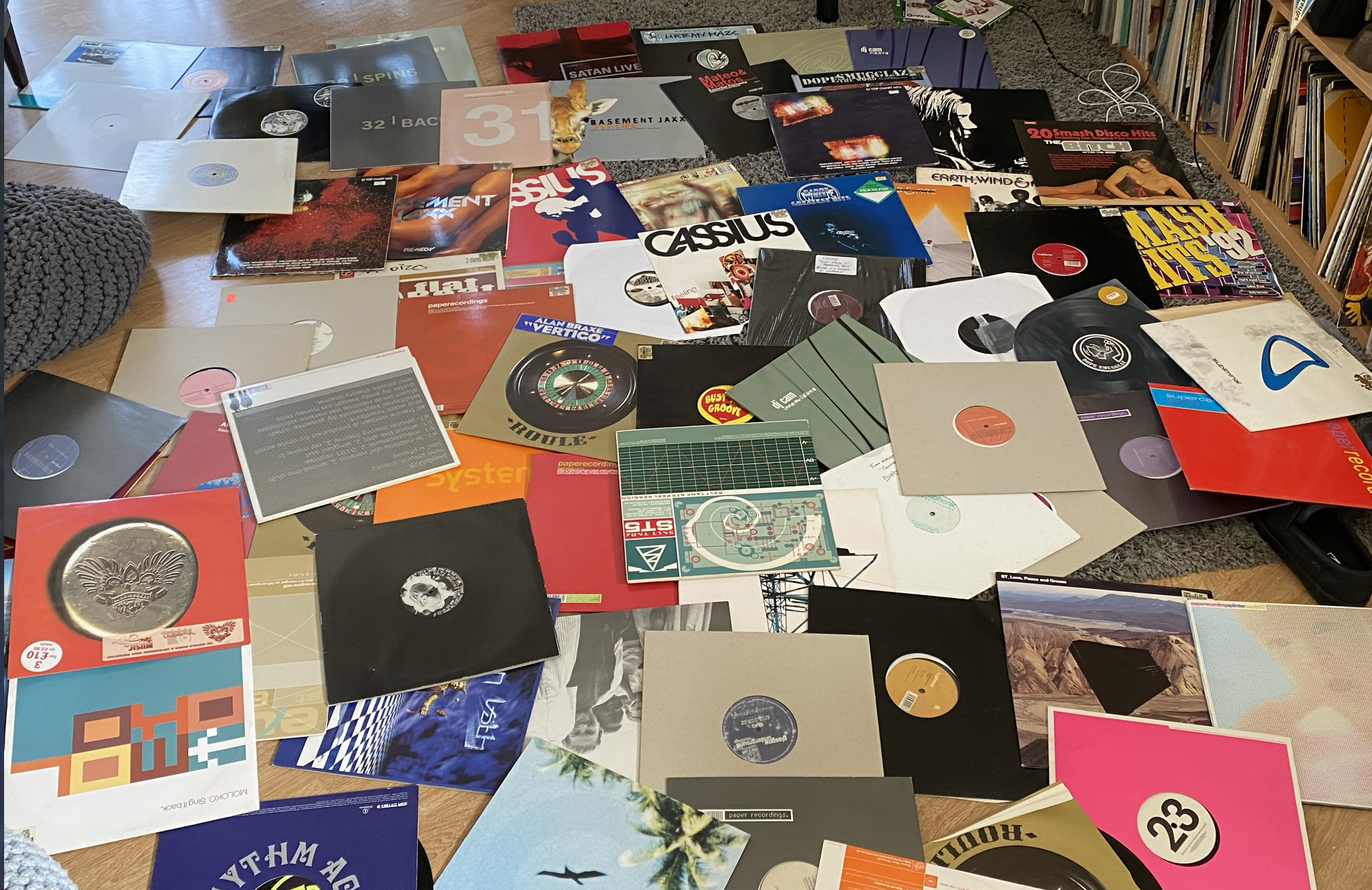 A collection of vinyl records spread out on a wooden table, with book shelves in the background and some electronic cords visible on the floor.