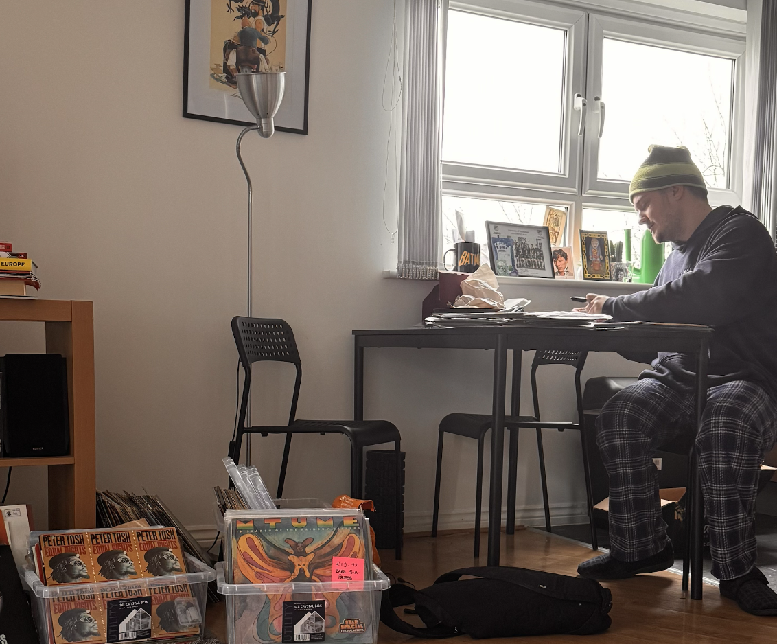 A man sitting at a desk in a room with a window, wearing a green beanie, glasses, a dark hoodie, and plaid pajama pants. There are framed pictures and a mug on the windowsill, and a pile of papers and items on the desk. A plastic container with comic books and Vinyl records is on the floor near the man, and a black backpack is next to them.