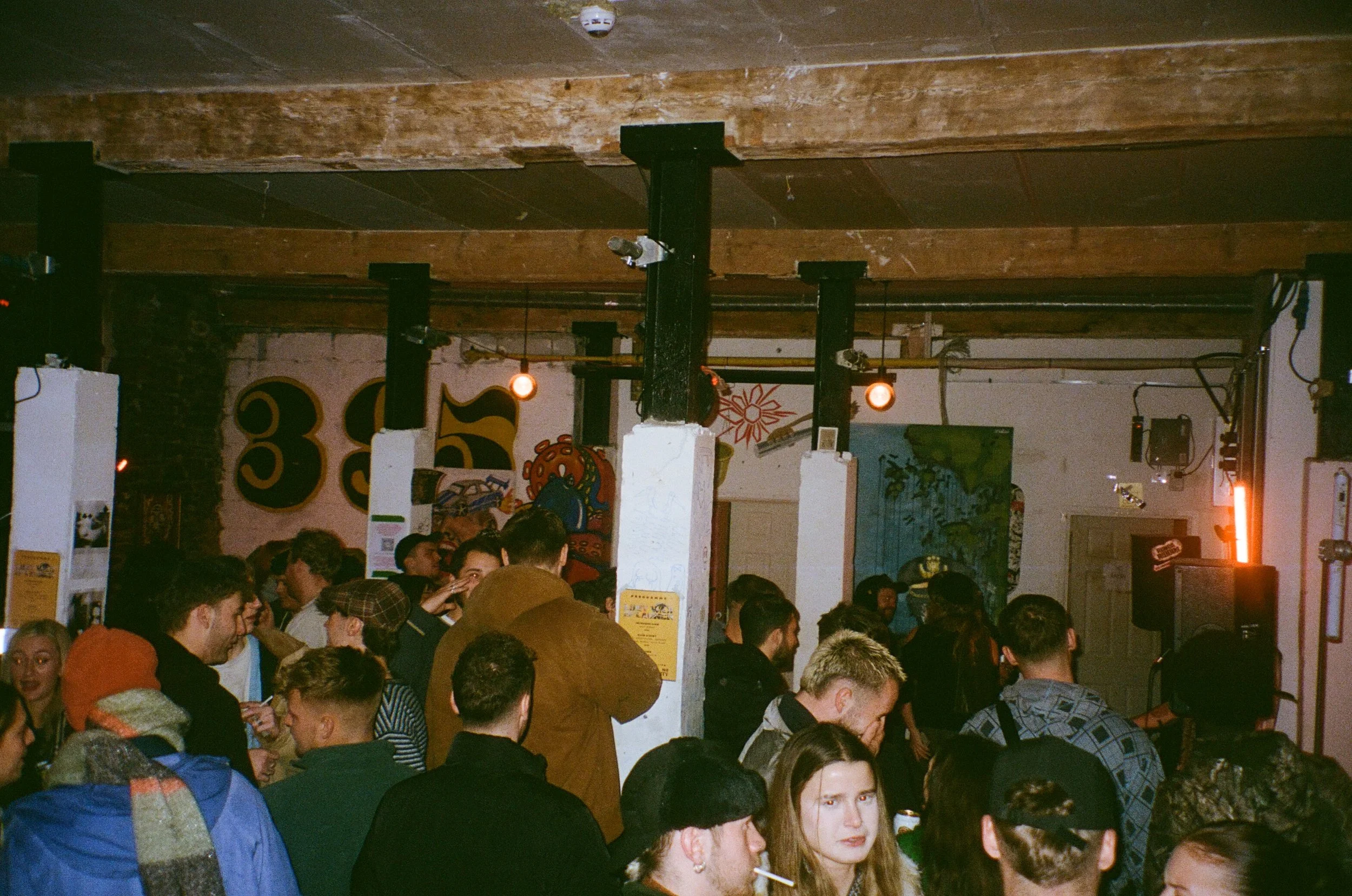 Crowd of people gathered indoors under a wooden ceiling with several hanging lights and visible security cameras.