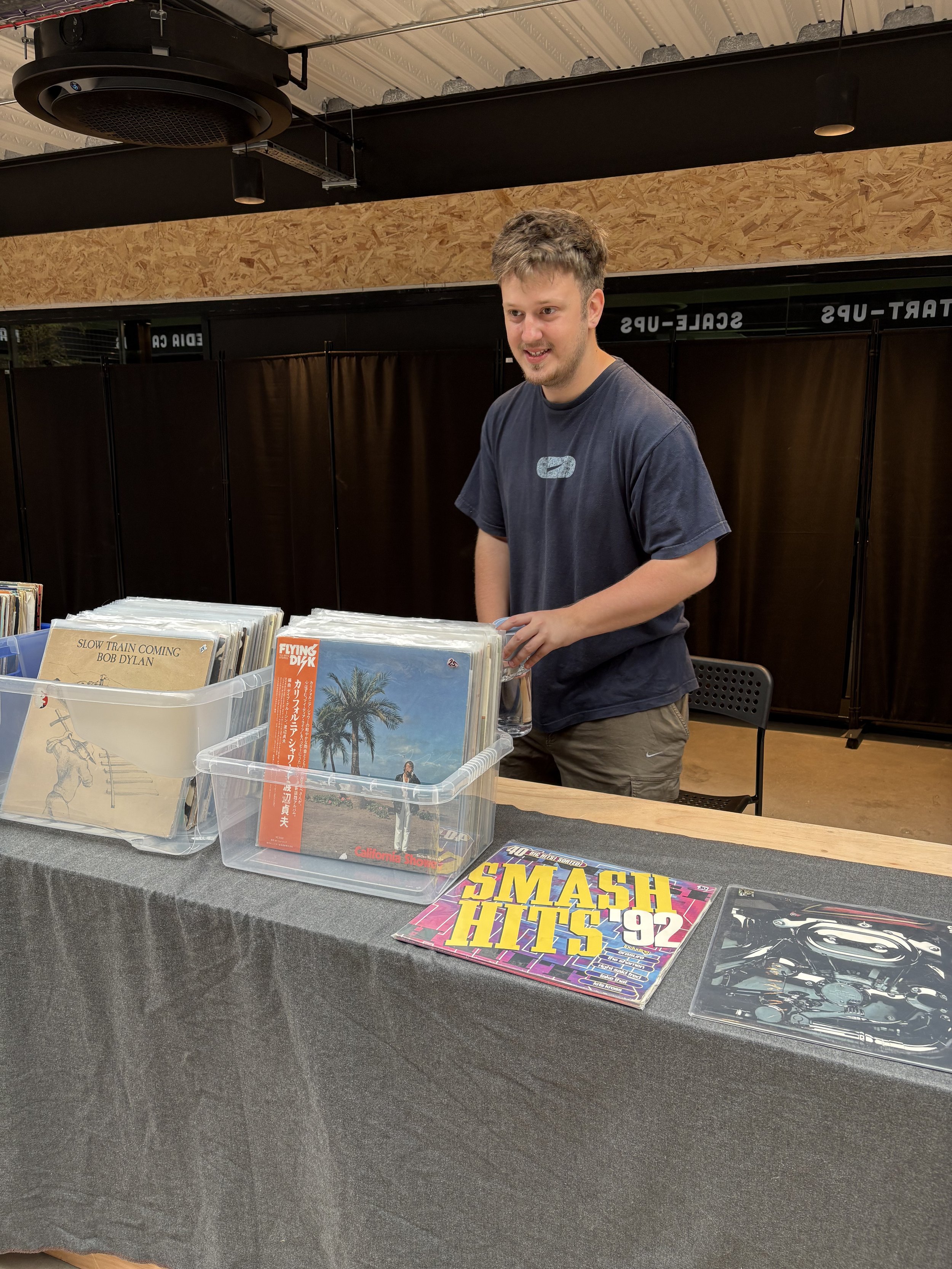 A person browsing through vinyl records at a music sale or store. There are several records on display, including 'Slow Train Coming' by Bob Dylan and 'Smash Hits 92'. The background features a black divider and a wooden wall, with part of a ceiling and lighting visible.