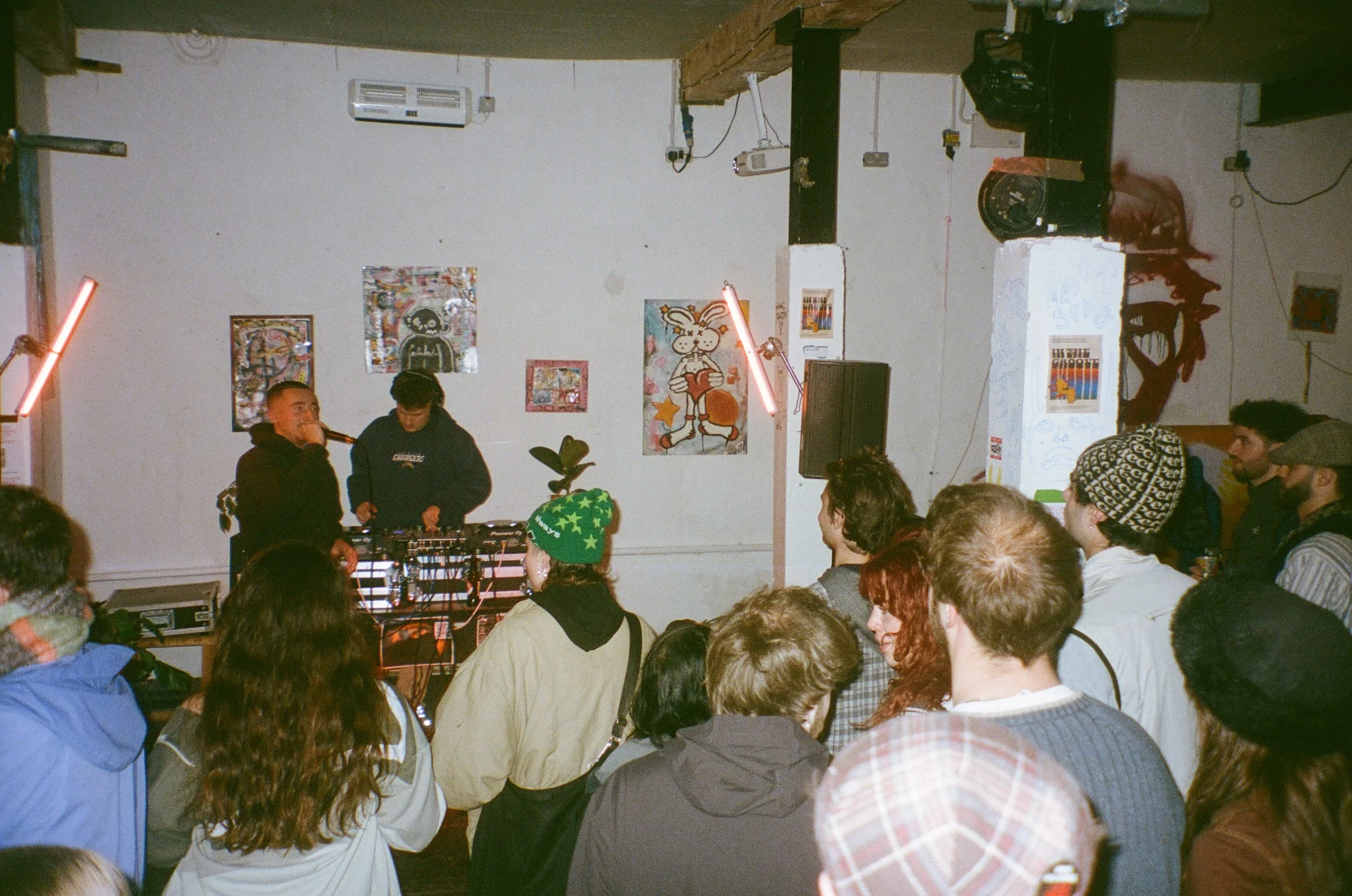 A group of people gathered in a room with artworks on the wall, where two DJs are performing with music equipment, illuminated by pink neon lights.