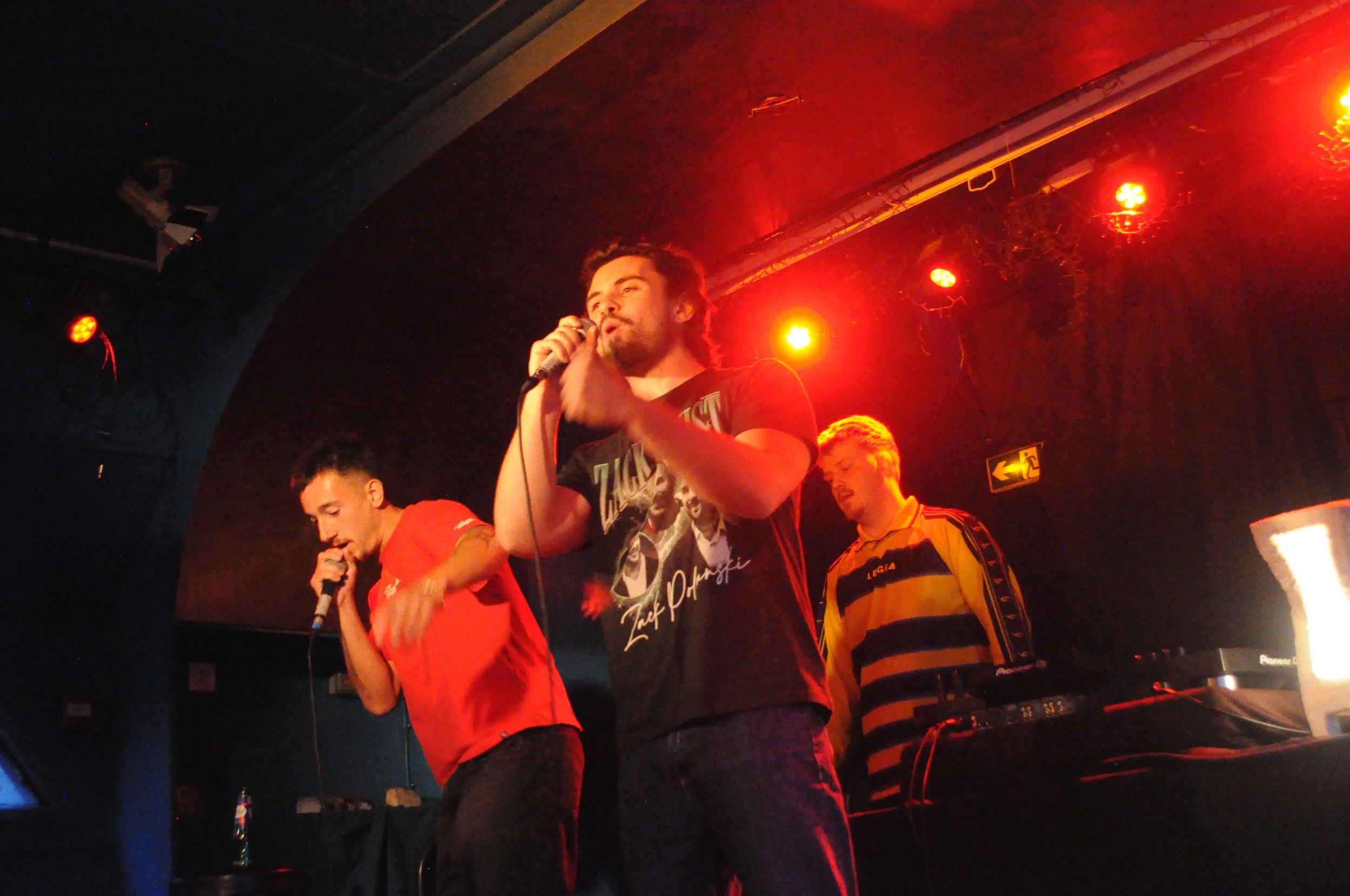 Three men on stage singing into microphones under red stage lights, with DJ equipment nearby.