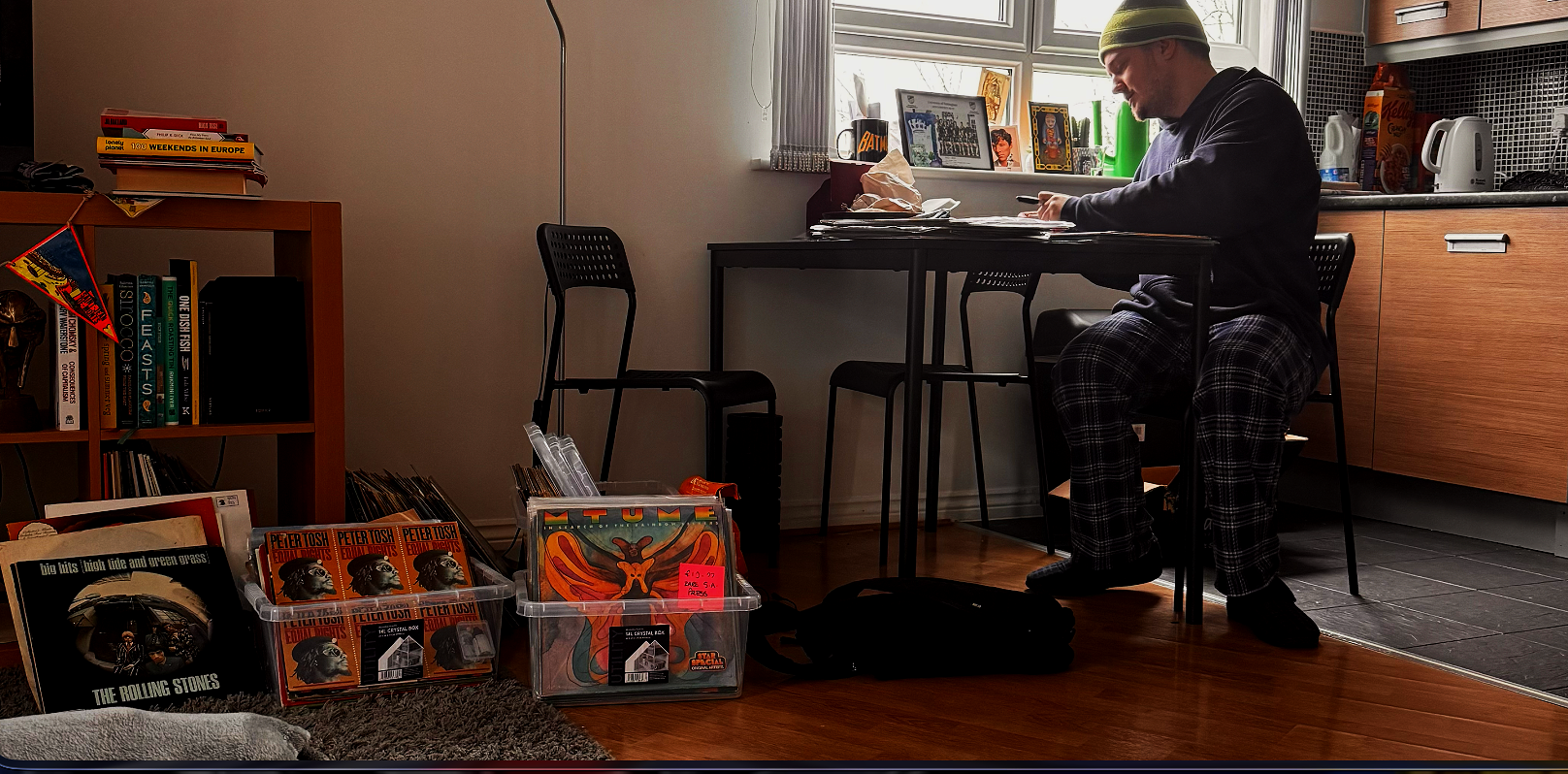 A man sitting at a dining table using a smartphone in a kitchen, with albums and books on the floor and shelf nearby, and a window with items on its sill in the background.