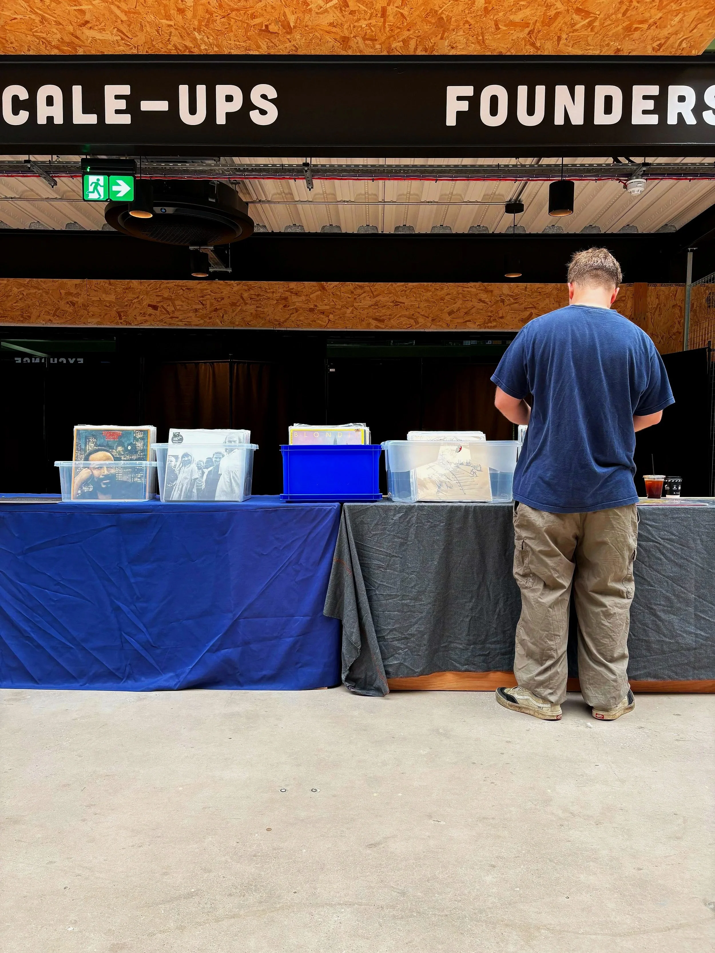 A person wearing a blue T-shirt and beige pants standing in front of a table with vinyl records displayed in plastic bins at an indoor event with a sign that reads 'SCALE-UPS FOUNDERS' and a green emergency exit sign above.