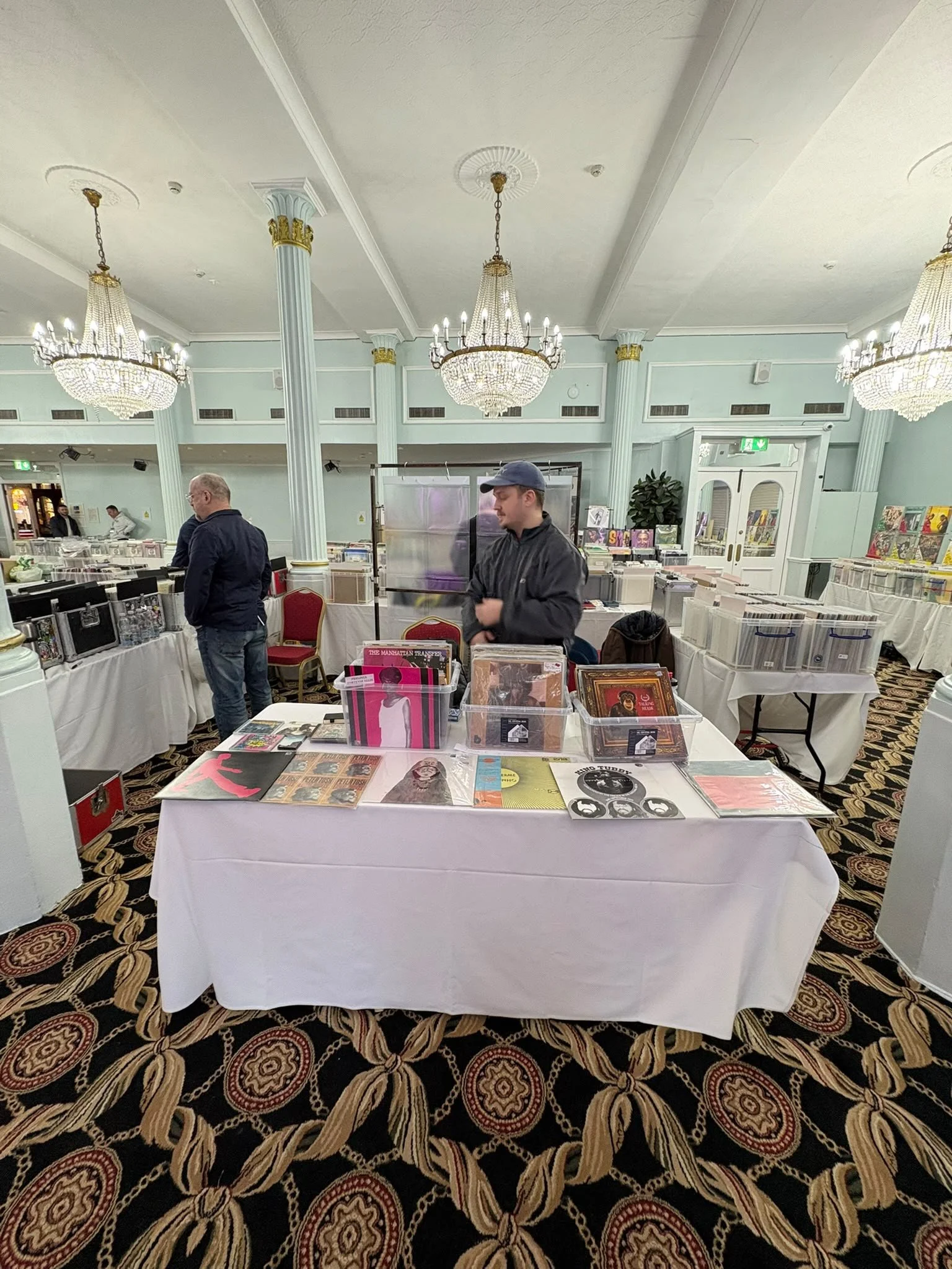 A view of an indoor event space with elegant chandeliers, tall columns, and tables covered with white cloths displaying vinyl records and music-related items. People are browsing the tables, and there are display racks in the background.