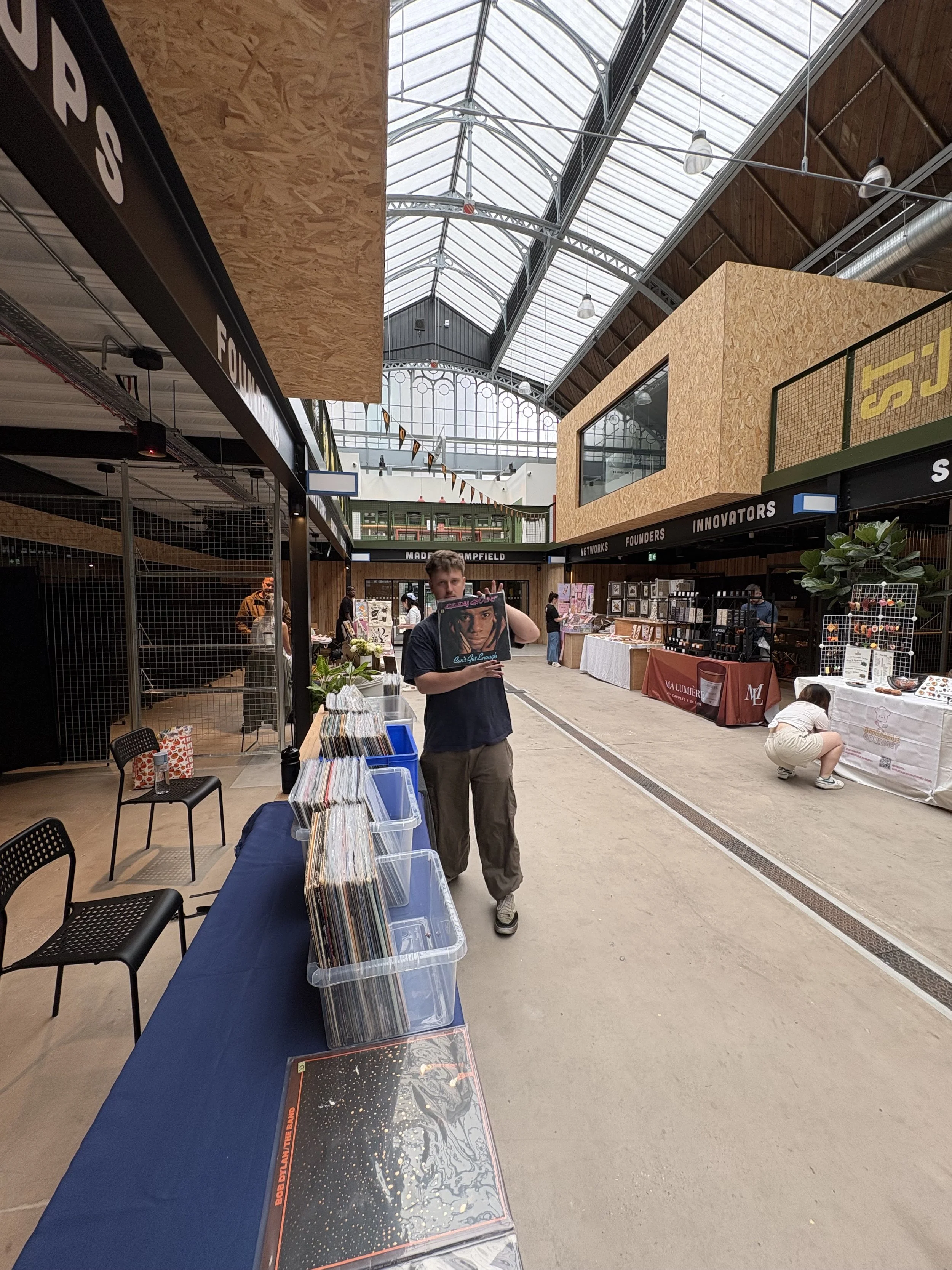 A person holding a vinyl record in front of a display of records at an indoor market or venue, with vendor booths and high arched glass ceiling.