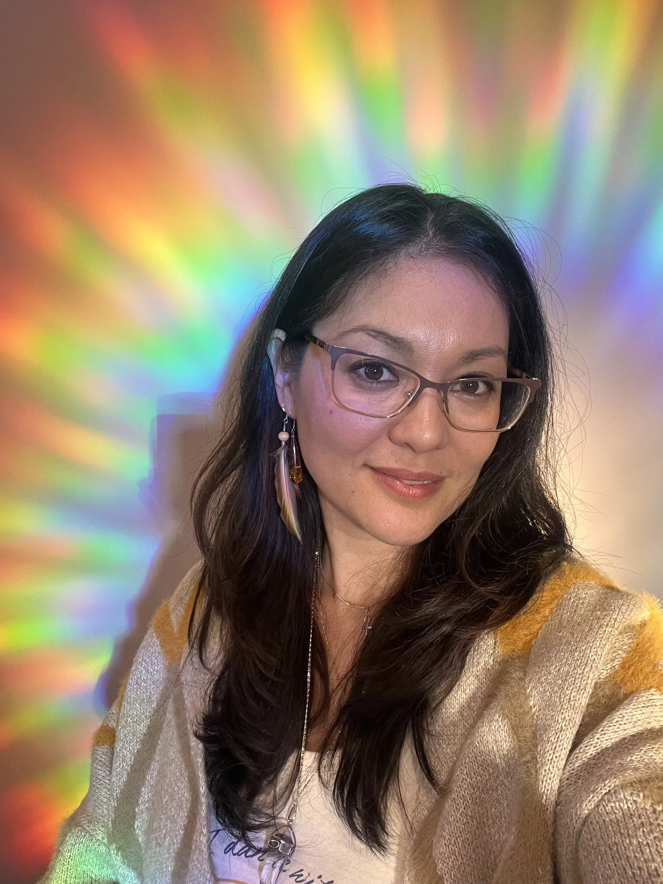 A woman with long dark hair, glasses, earrings, and a nose ring, smiling at the camera. She is wearing a beige sweater and a white shirt with a graphic. The background features colorful rainbow-like light reflections.