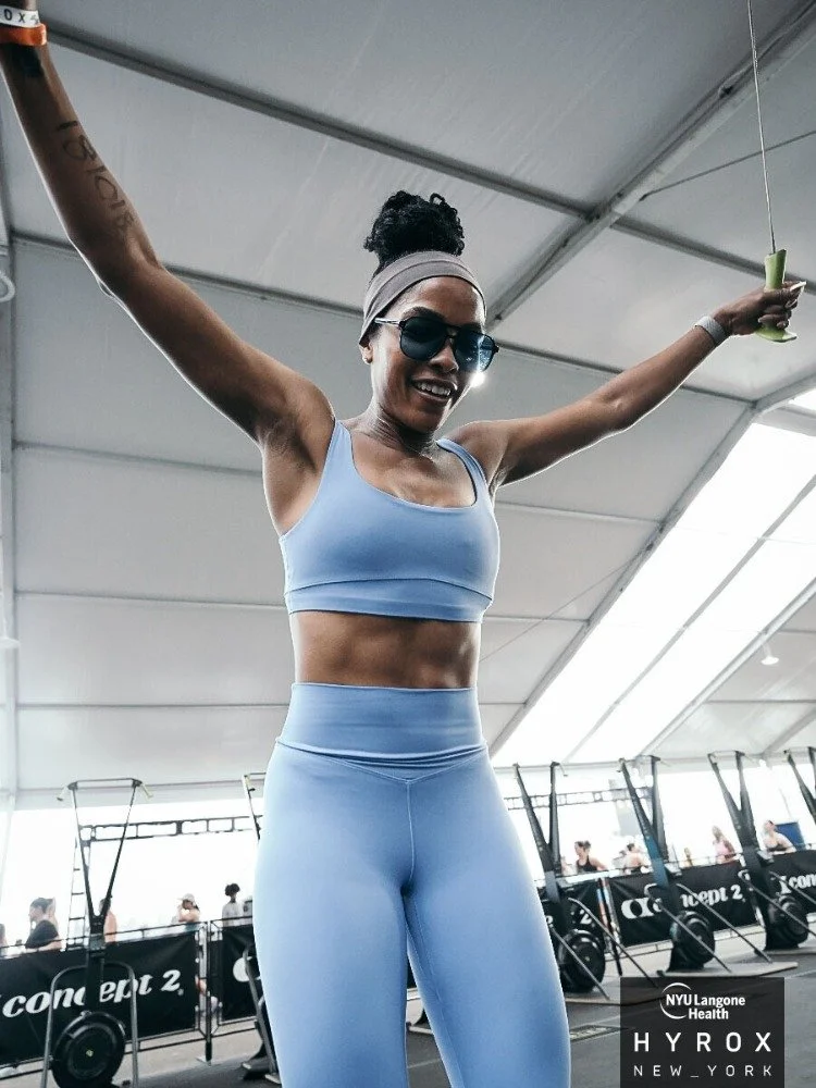 A woman in workout clothes smiling with arms raised inside a gym.