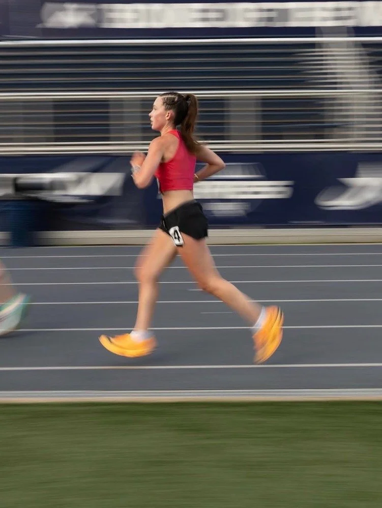 A female runner in a red tank top and black shorts running on a track with a blurred blue bus in the background.