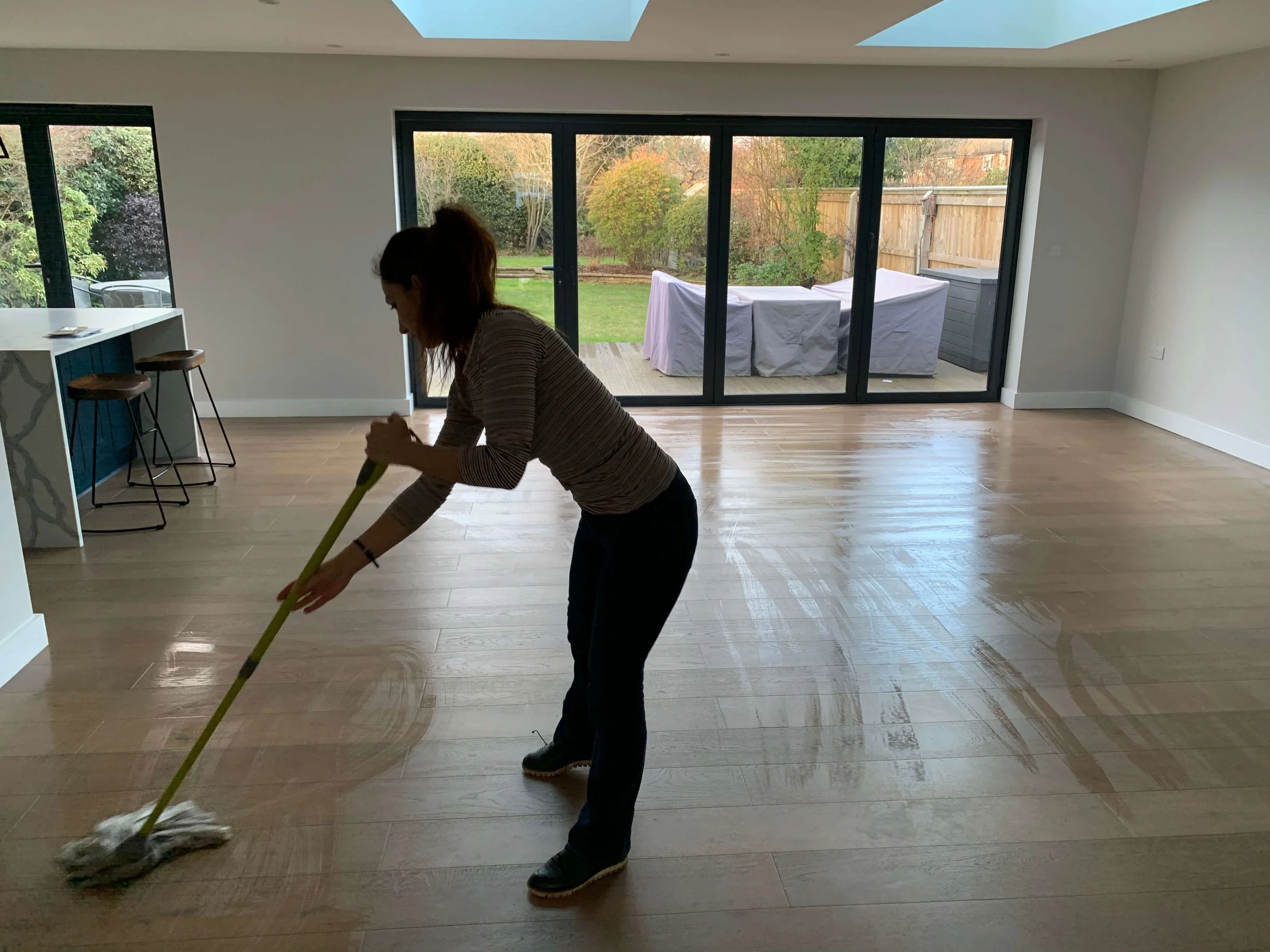 A person sweeping hardwood flooring inside a modern, bright living space with large sliding glass doors leading to a backyard with trees and a wooden fence.