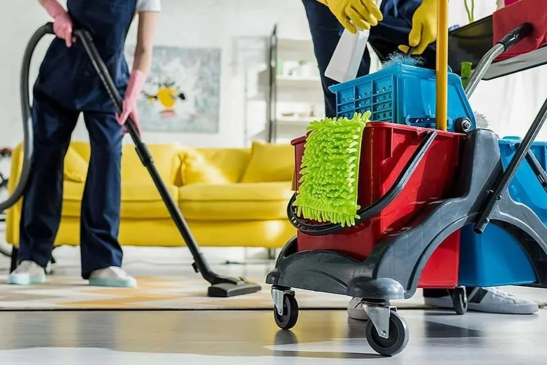 A person cleaning a floor with a vacuum cleaner in a living room with a yellow sofa and colorful cleaning cart in the foreground.