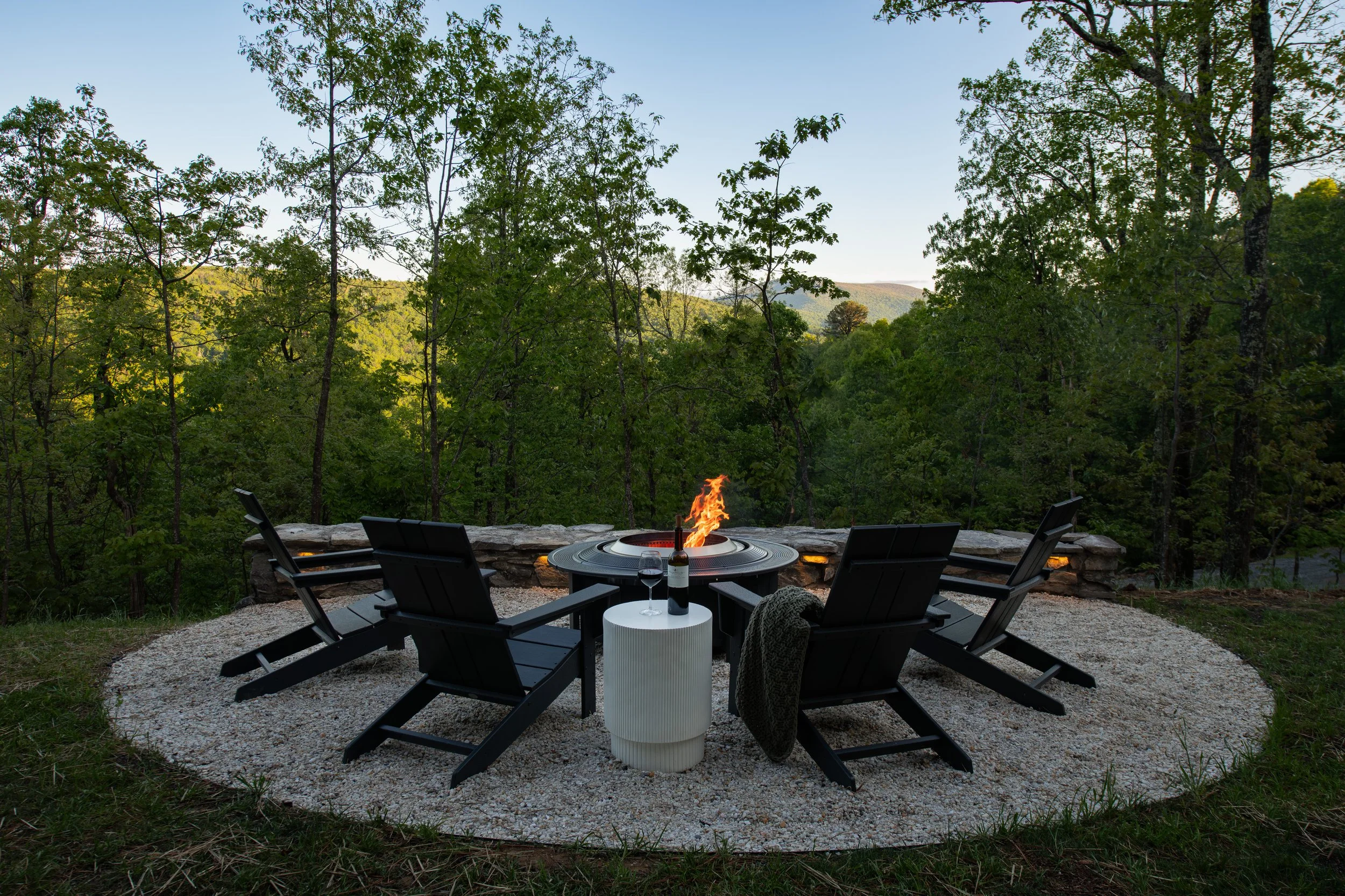 An outdoor patio with a round fire pit table, surrounding black chairs, a white side table with a wine glass and bottle, and green trees in the background during evening.