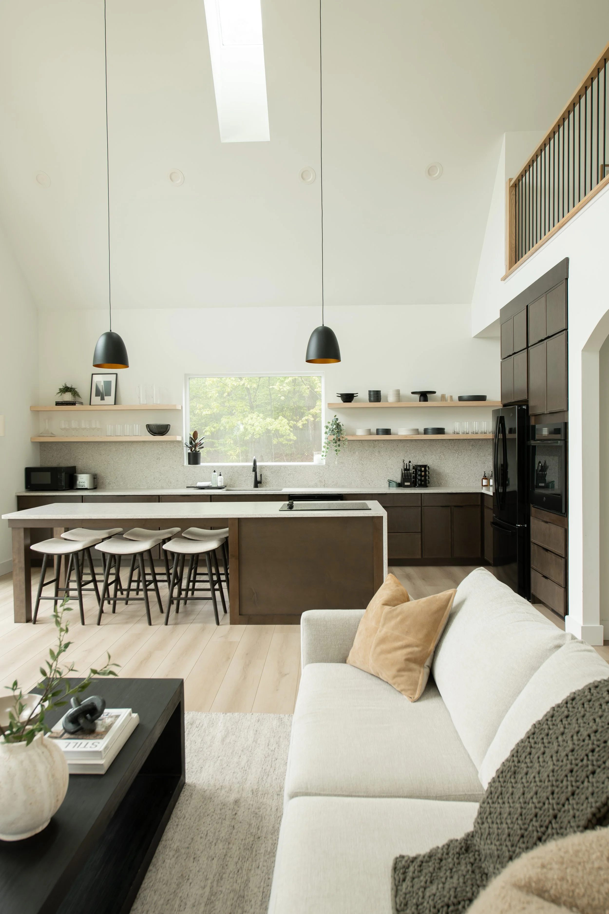 A modern living room and kitchen with light walls, a white couch with beige and gray pillows, a black coffee table with a beige vase and books, black pendant lights, open shelves with dishes, a large window showing green trees, and black kitchen appl