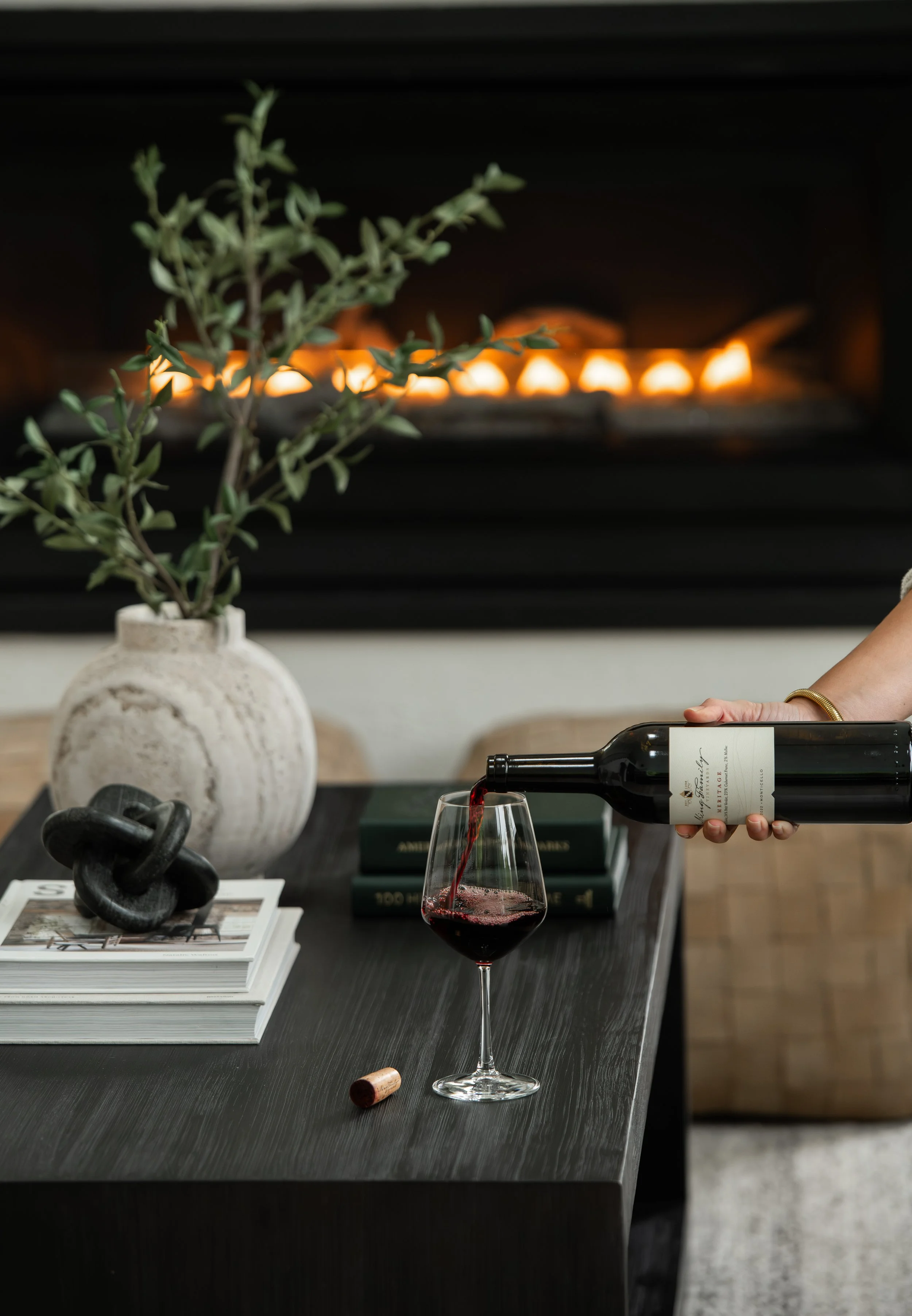A person pouring red wine into a glass on a dark wooden table. In the background, there is a decorative vase with green leafy branches, a black abstract sculpture, and a stack of books. The scene is set in a modern, stylish interior with a black fire