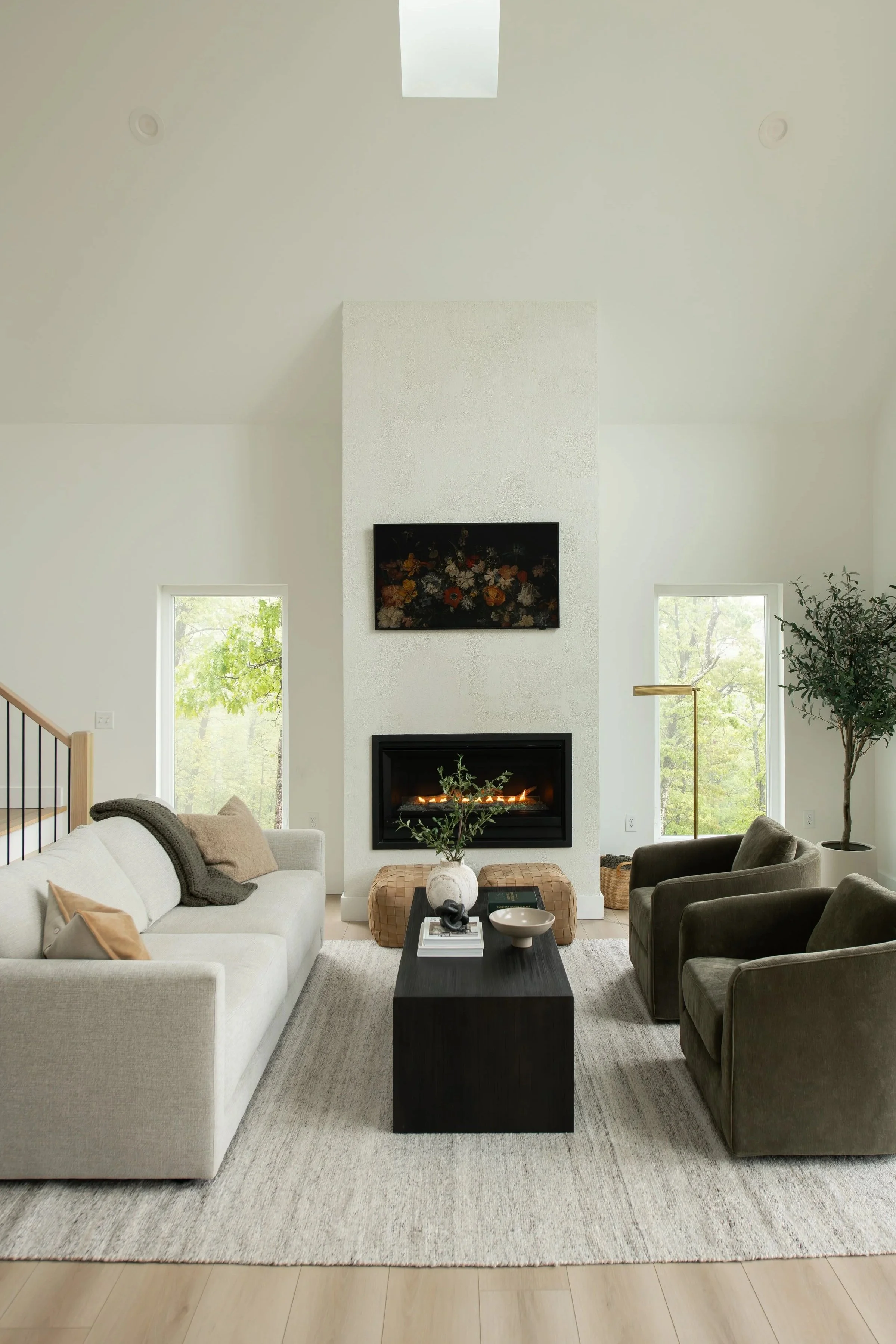 Modern living room with a white sofa, black armchair, and a fireplace on a textured beige rug. Window views of green trees, a black coffee table with books and decor, and a tall green plant in the corner.