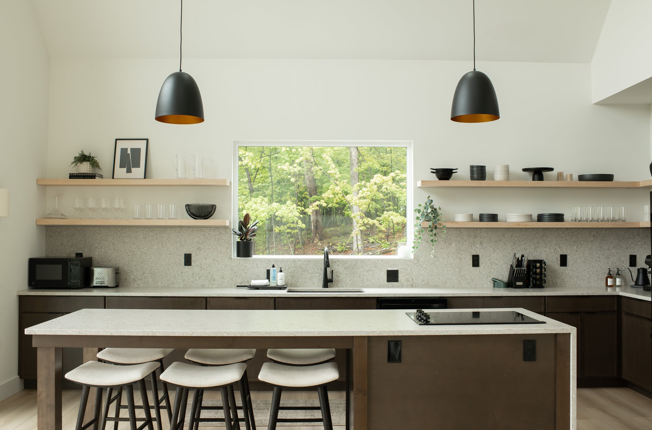 Modern kitchen with large window showing green trees outside, black pendant lights, open floating shelves with dishes and decorative items, island with four white chairs, and various kitchen appliances.