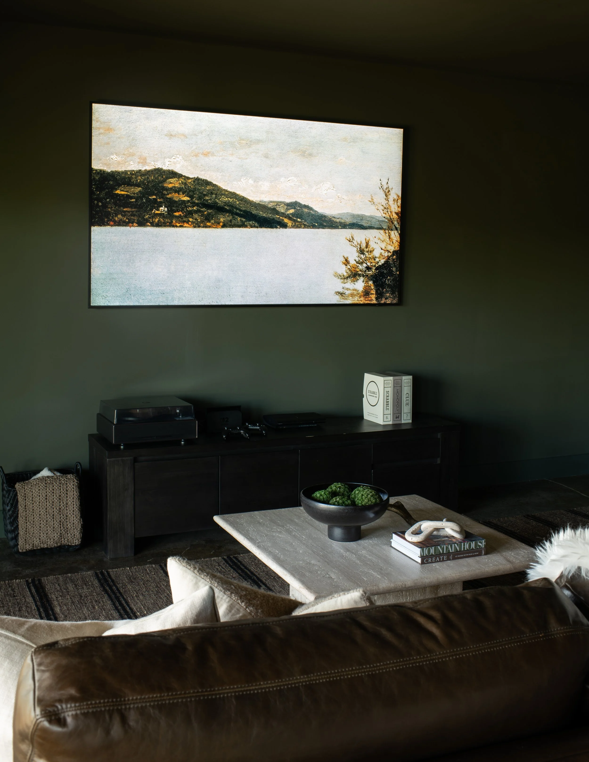 Living room with a large wall-mounted painting of a lakeside landscape, a dark wood cabinet with audio equipment, a low coffee table with a black bowl of green decorative balls, books, and a beige sofa.