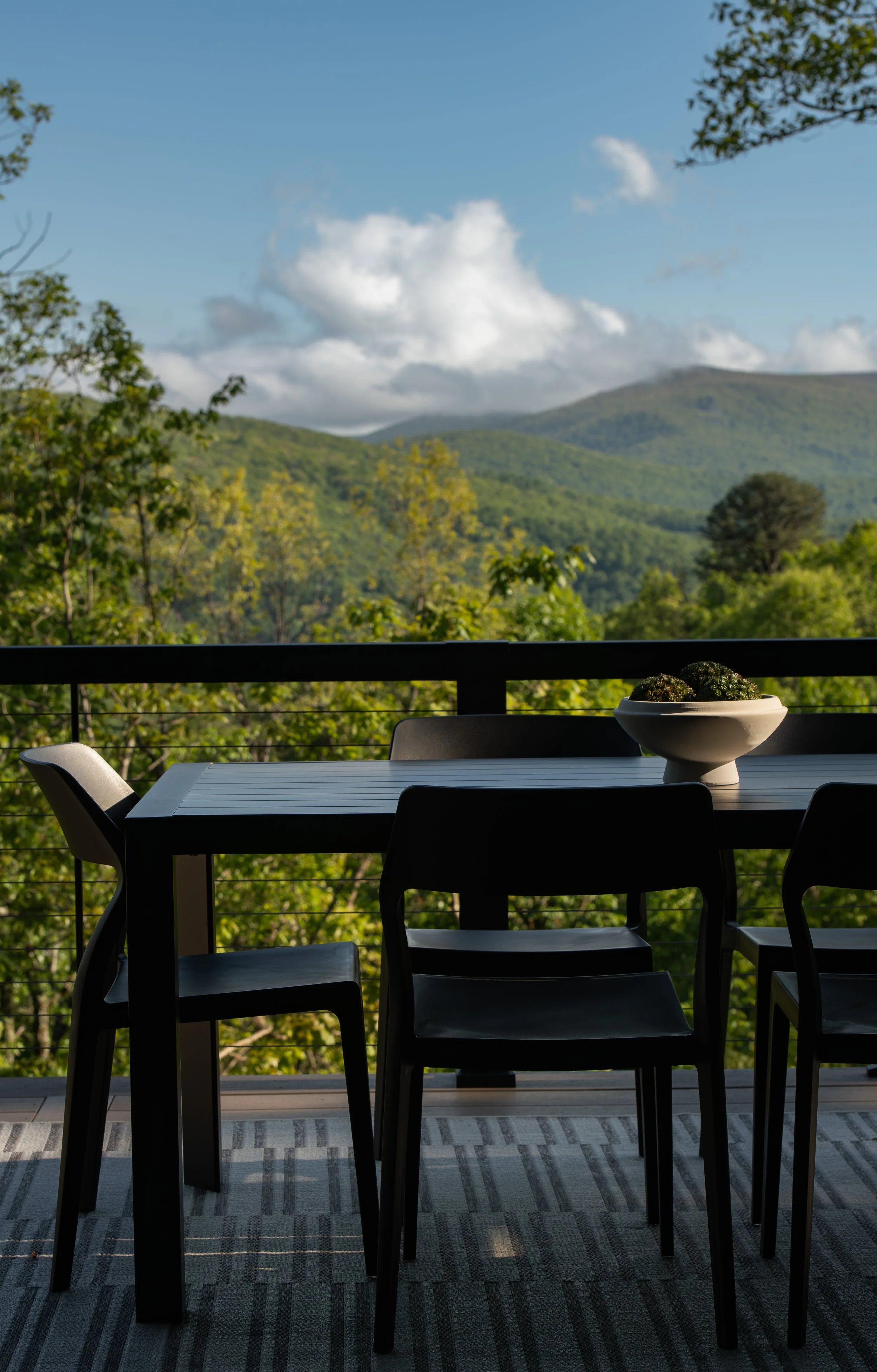 Outdoor dining area on a balcony with a mountain view of lush green trees and hills under a blue sky with white clouds. A large white bowl with moss balls sits on a black rectangular table with black chairs.