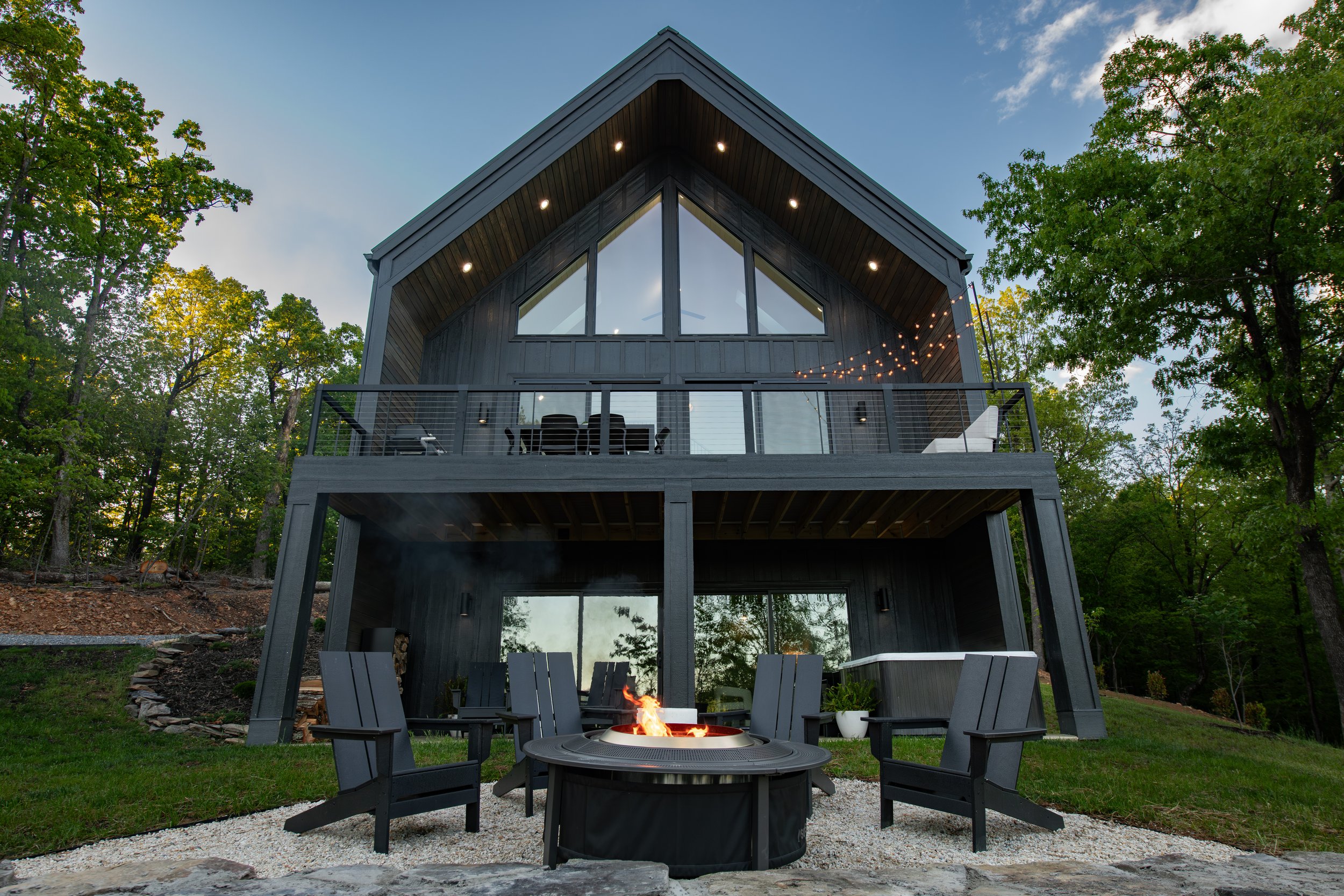 Modern two-story house with a balcony and large glass windows, surrounded by trees, with an outdoor firepit and chairs in the yard.