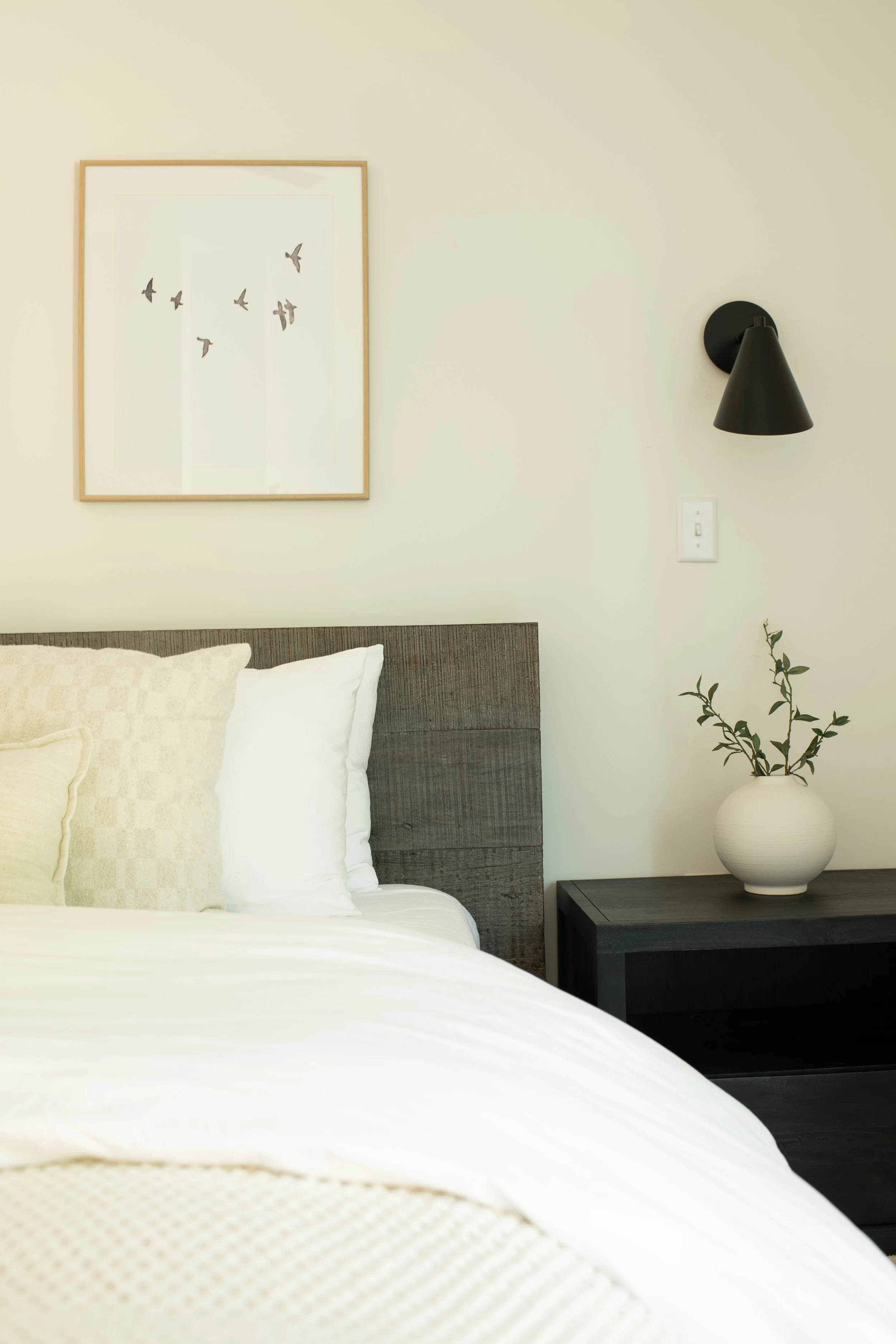 A bedroom with a bed, white sheets, and pillows. There is a bedside table with a white vase containing branches. A black wall-mounted lamp and a framed picture of birds flying on the wall.