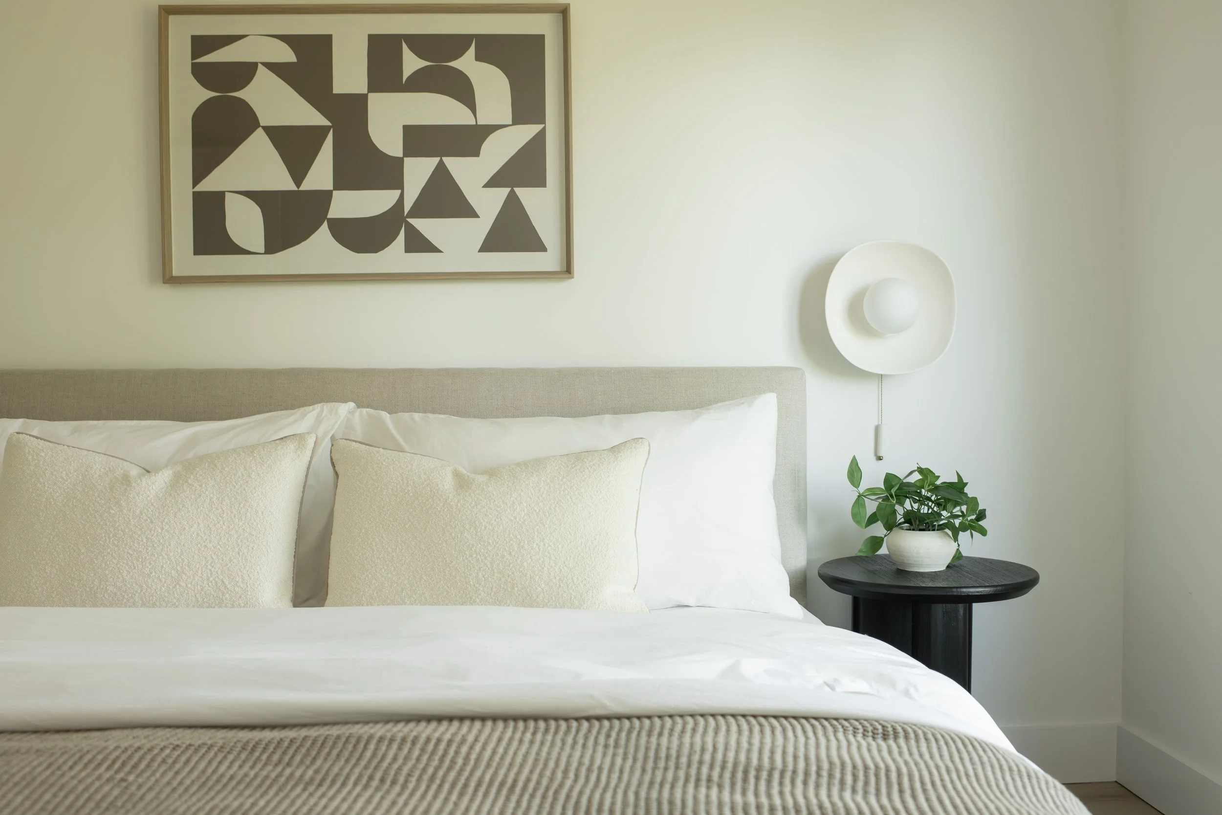 A neatly made bed with cream-colored pillows and bedding. A black side table with a potted plant on it stands beside the bed. Above the bed, there is framed abstract art with geometric shapes in shades of gray and black, and a white wall-mounted ligh