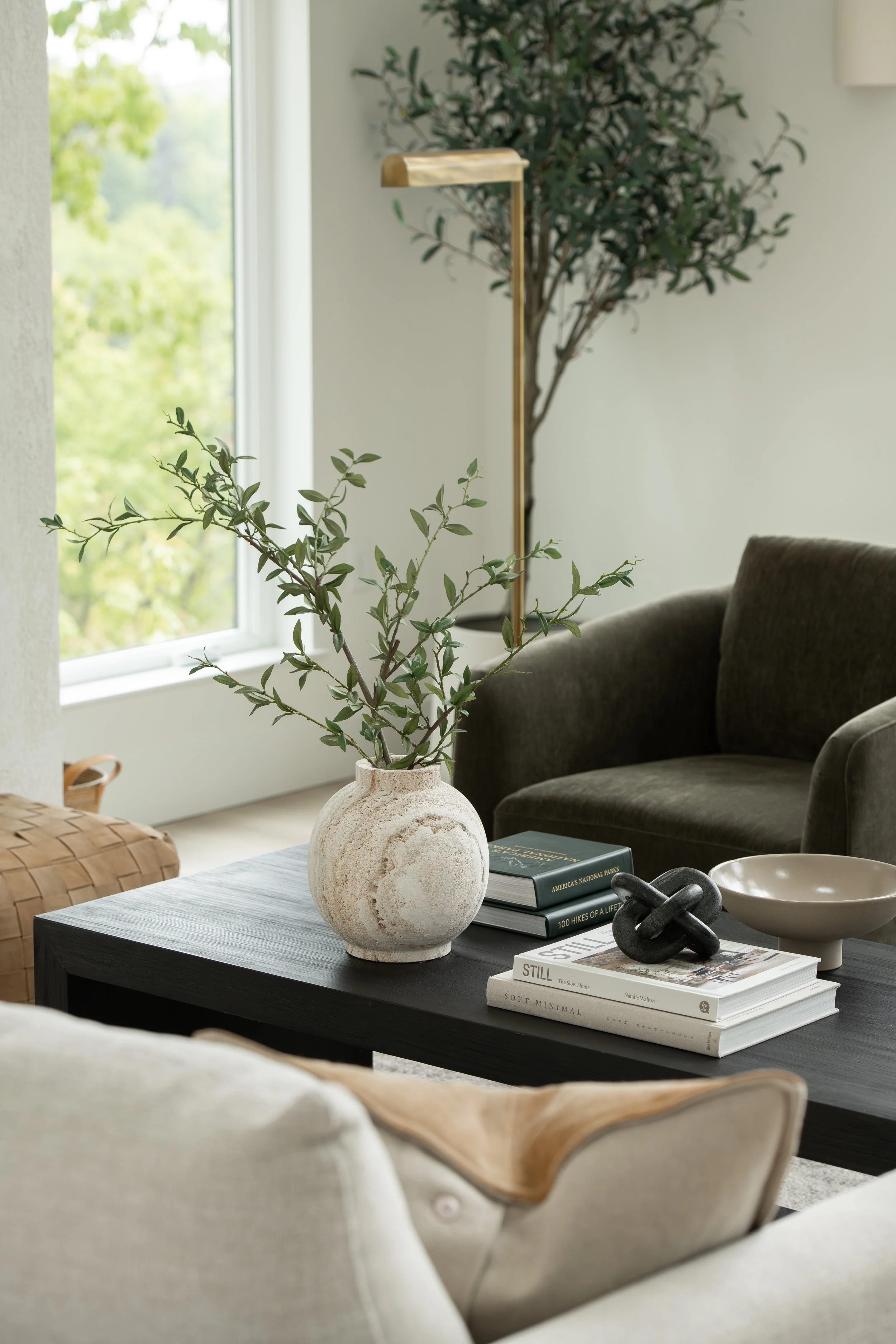 Living room interior with a black coffee table, a beige armchair, a ceramic vase with green foliage, stacked books, and a window with green trees outside.