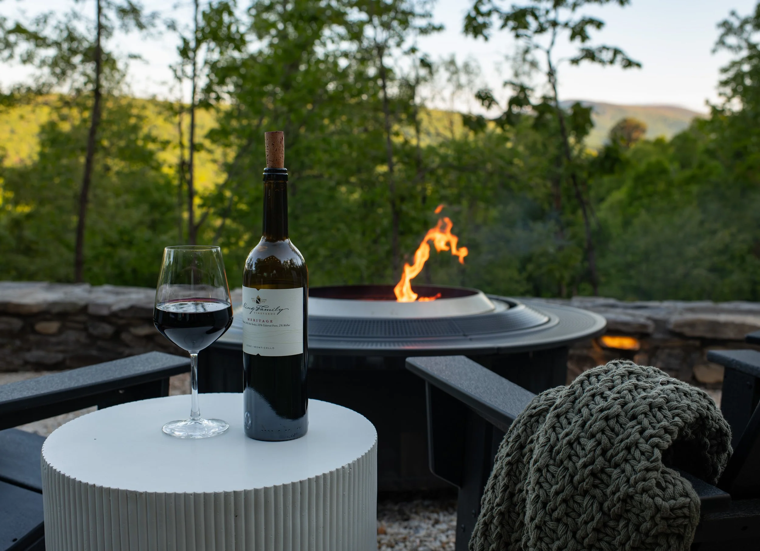 A glass of red wine and a wine bottle on a white round table outdoors with a fire pit and a scenic view of trees and hills in the background.