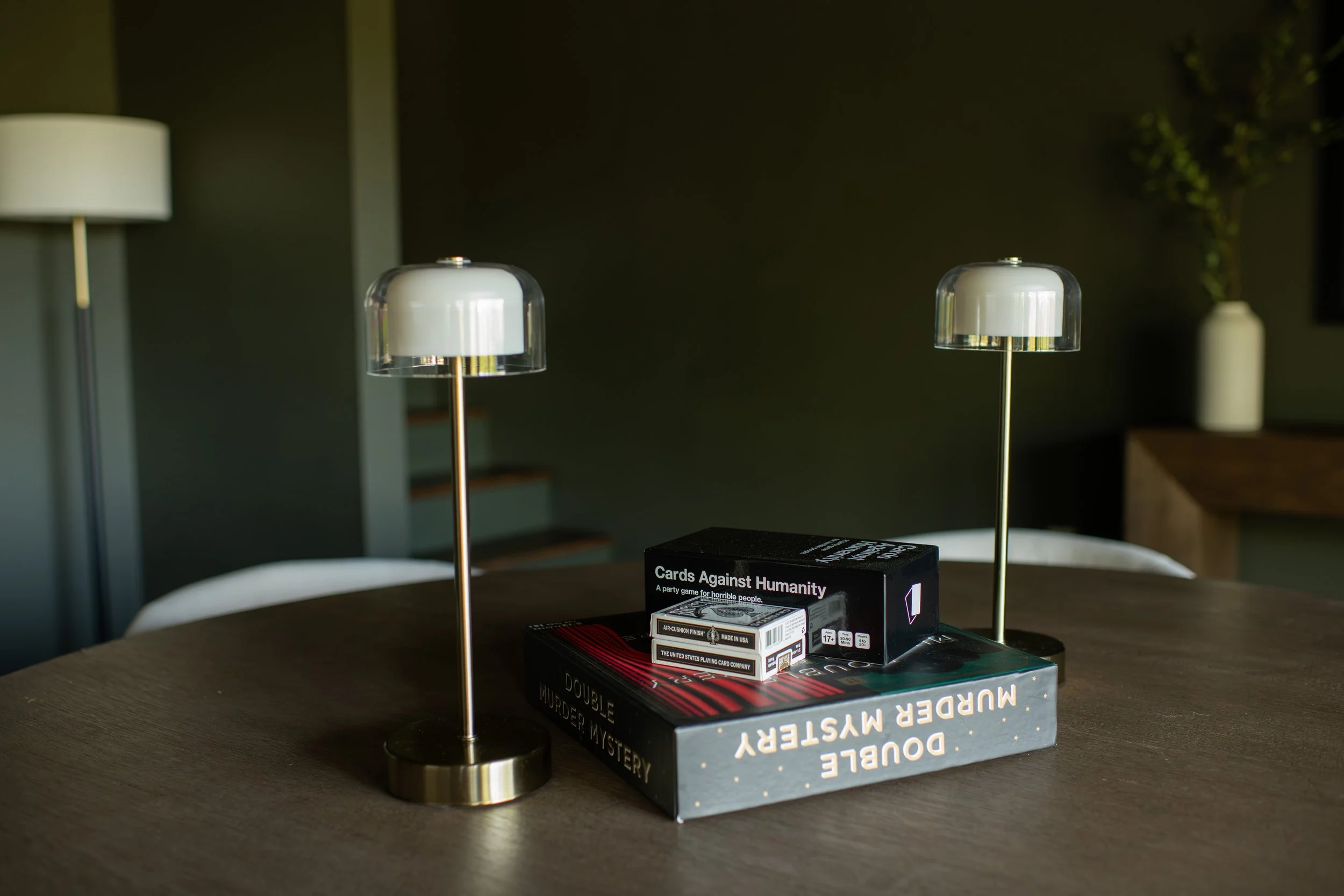 Two modern table lamps with white and transparent shades on a wooden table, with two stacked books and a card game box titled 'Cards Against Humanity' in the foreground, and a blurred background including a floor lamp and a vase with green plants.