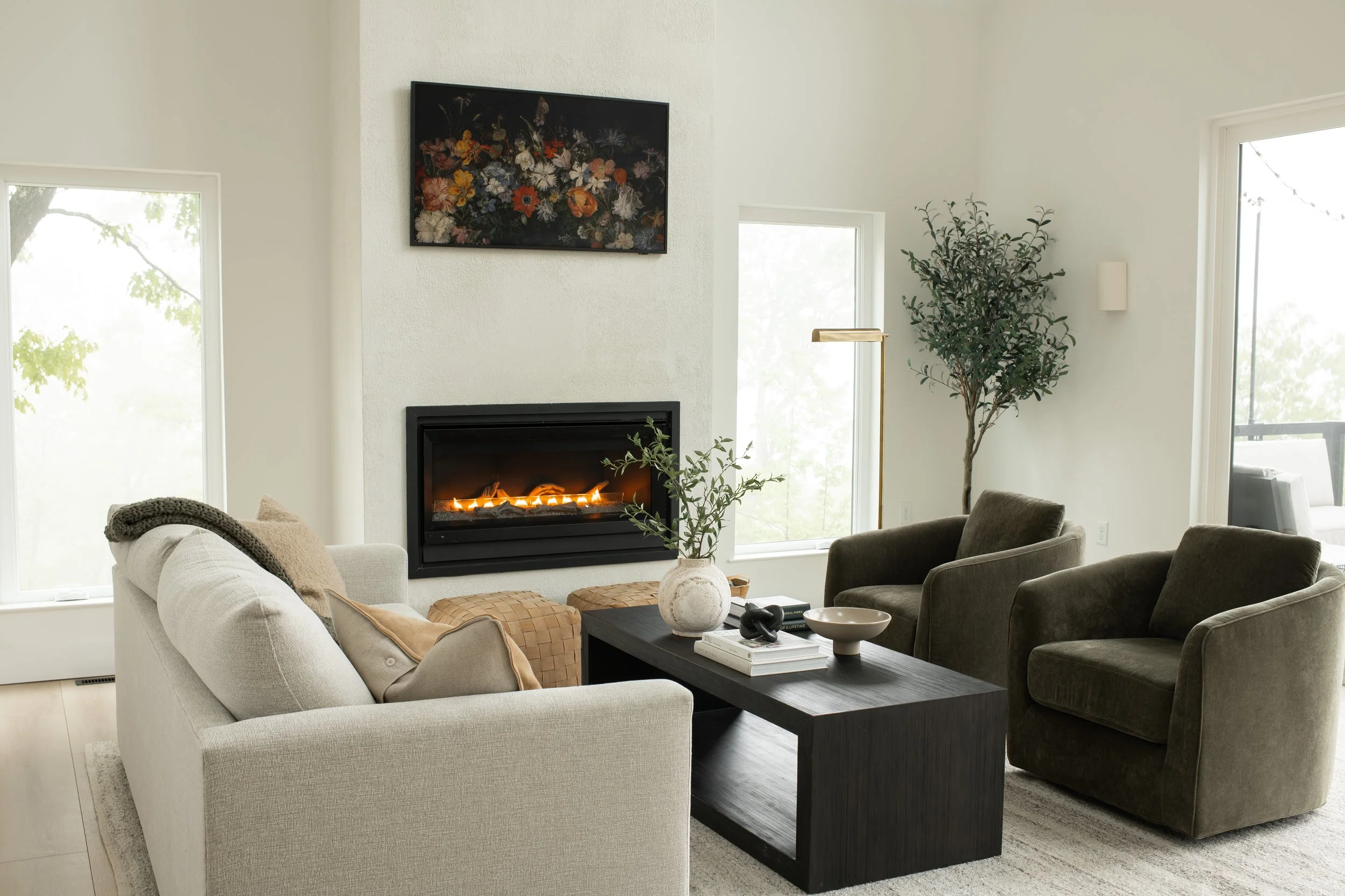 Living room with beige sofa, dark armchairs, a black coffee table, a large potted plant, a wall-mounted TV showing a floral image, and a fireplace with a fire, surrounded by large windows letting in natural light.