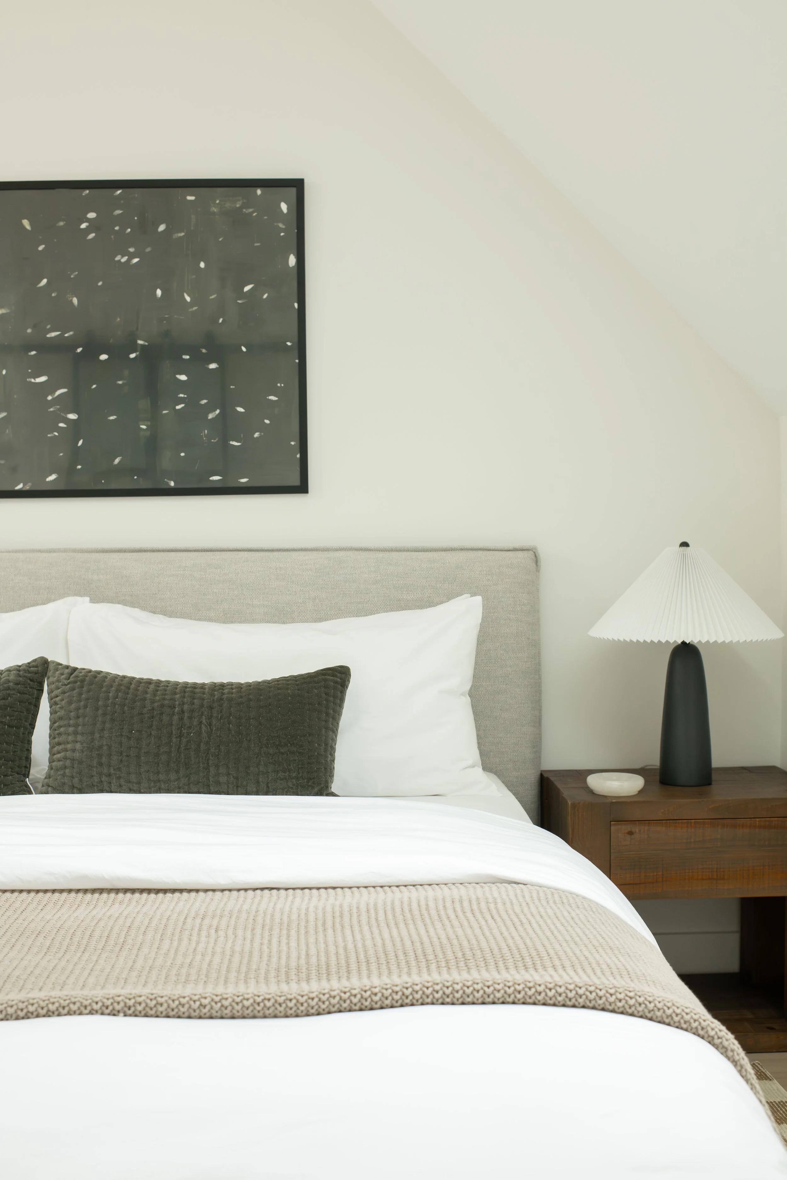 A bedroom with a bed, pillows, a nightstand, a lamp, and a wall art piece in a modern, minimalist style.