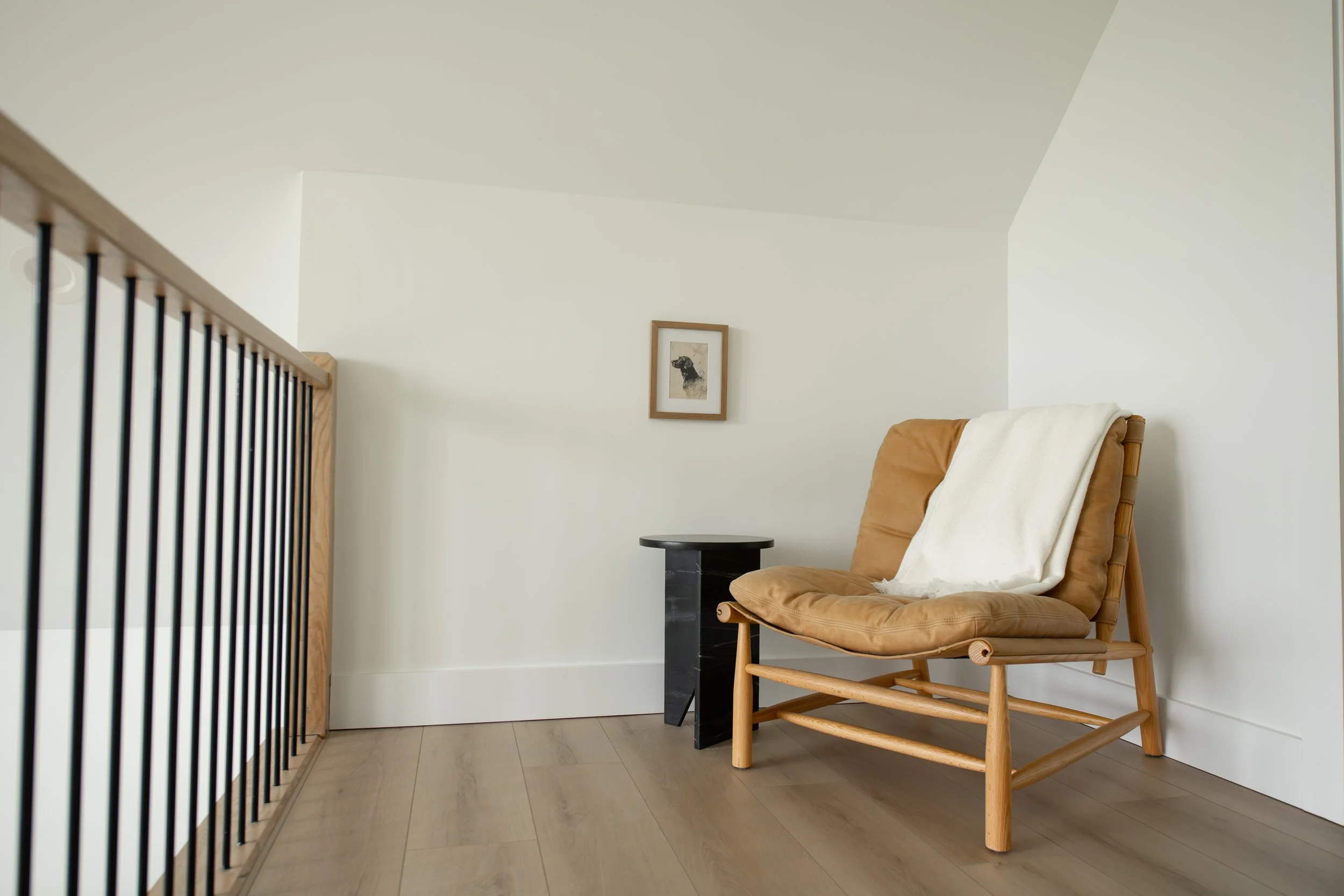 A cozy armchair with a blanket draped over it, placed next to a small black side table in a minimalistic, well-lit corner of a room. Framed artwork hangs on the white wall behind.