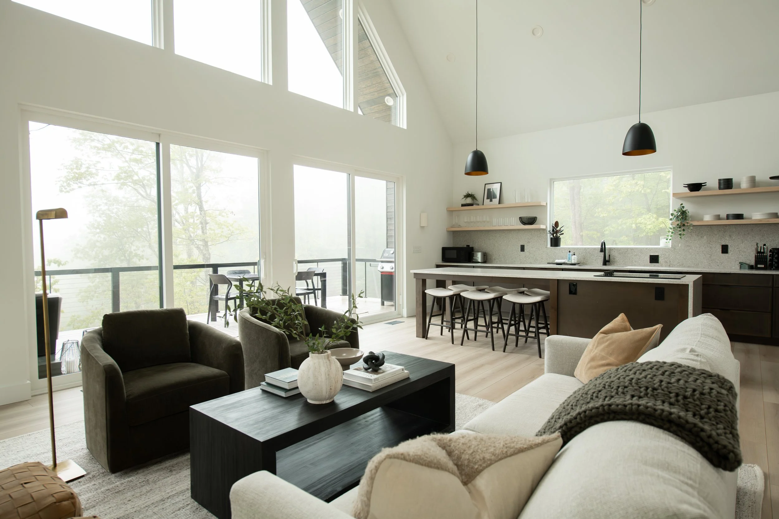 Open-concept living room and kitchen with large windows, modern furniture, a black coffee table, white and beige sofas, and a kitchen with open shelves, bar stools, and pendant lighting.