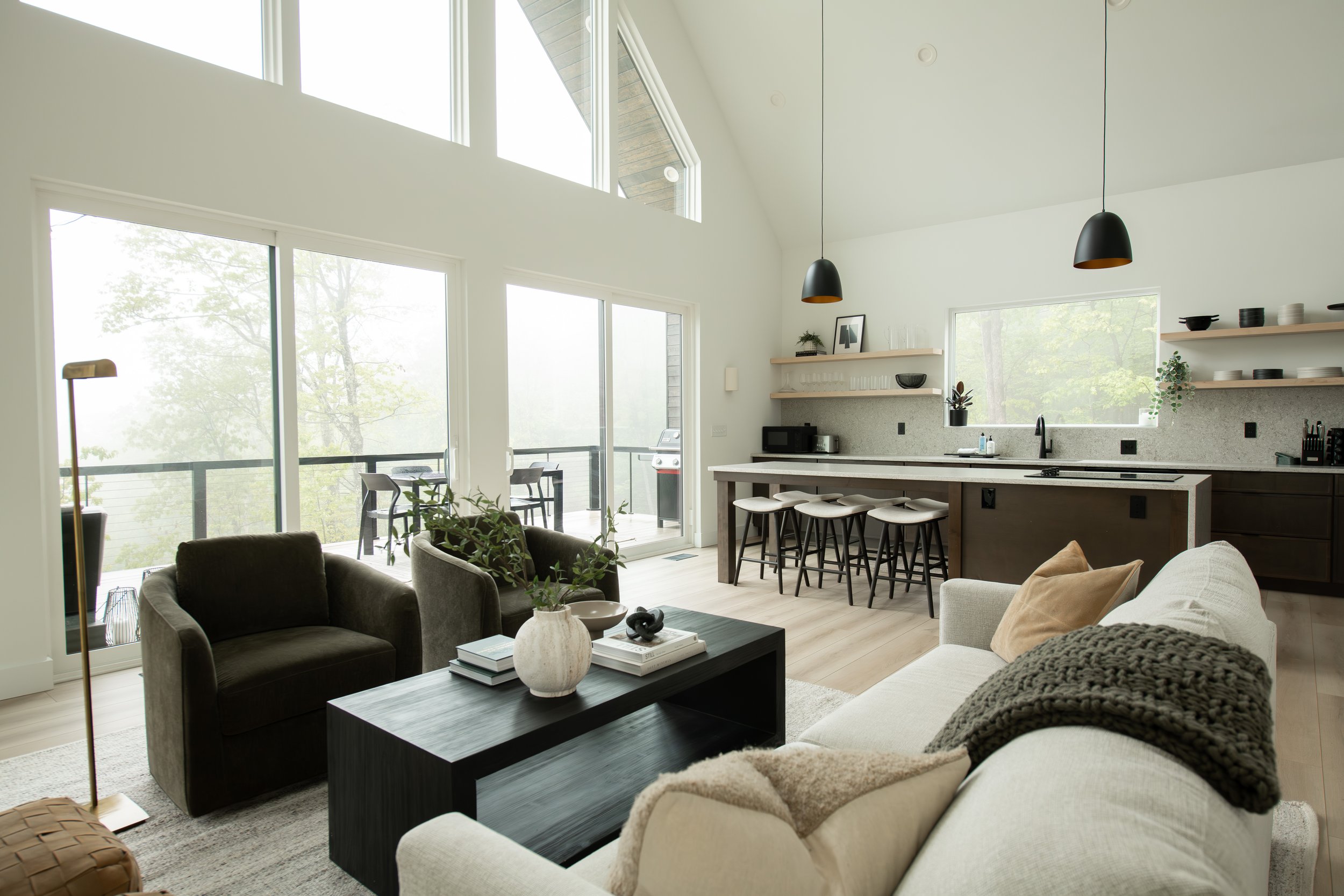Modern open-concept living room and kitchen with large windows, high ceiling, hanging pendant lights, and a balcony with outdoor furniture.